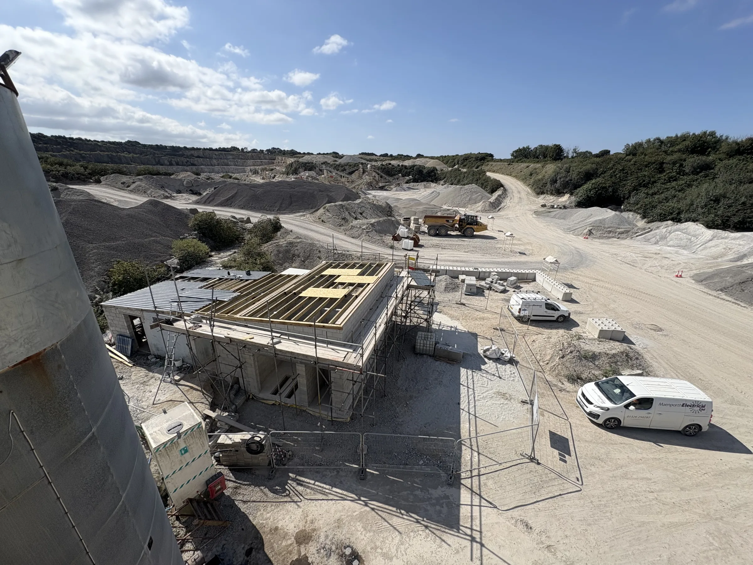 Elevated view of a scaffolded blockwork building with exposed timber roof framing, surrounded by white vans, aggregate piles and machinery.