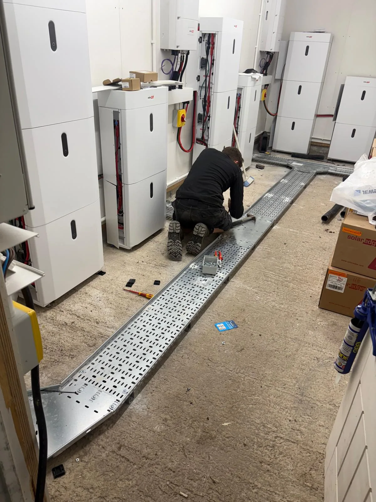 Worker kneeling beside a long perforated metal cable tray among white electrical units, exposed cables, tools and boxes.