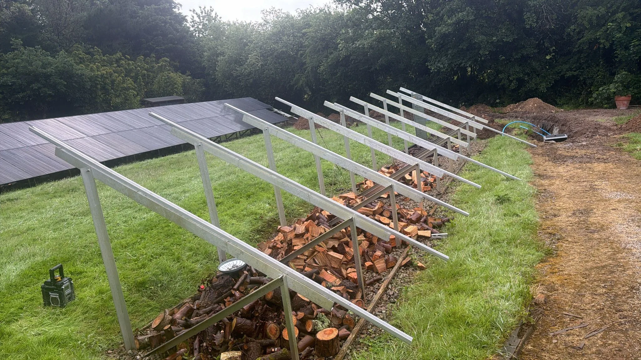 Rows of angled metal solar panel support frames on grass, with stacked cut logs beneath and panels visible to the left.