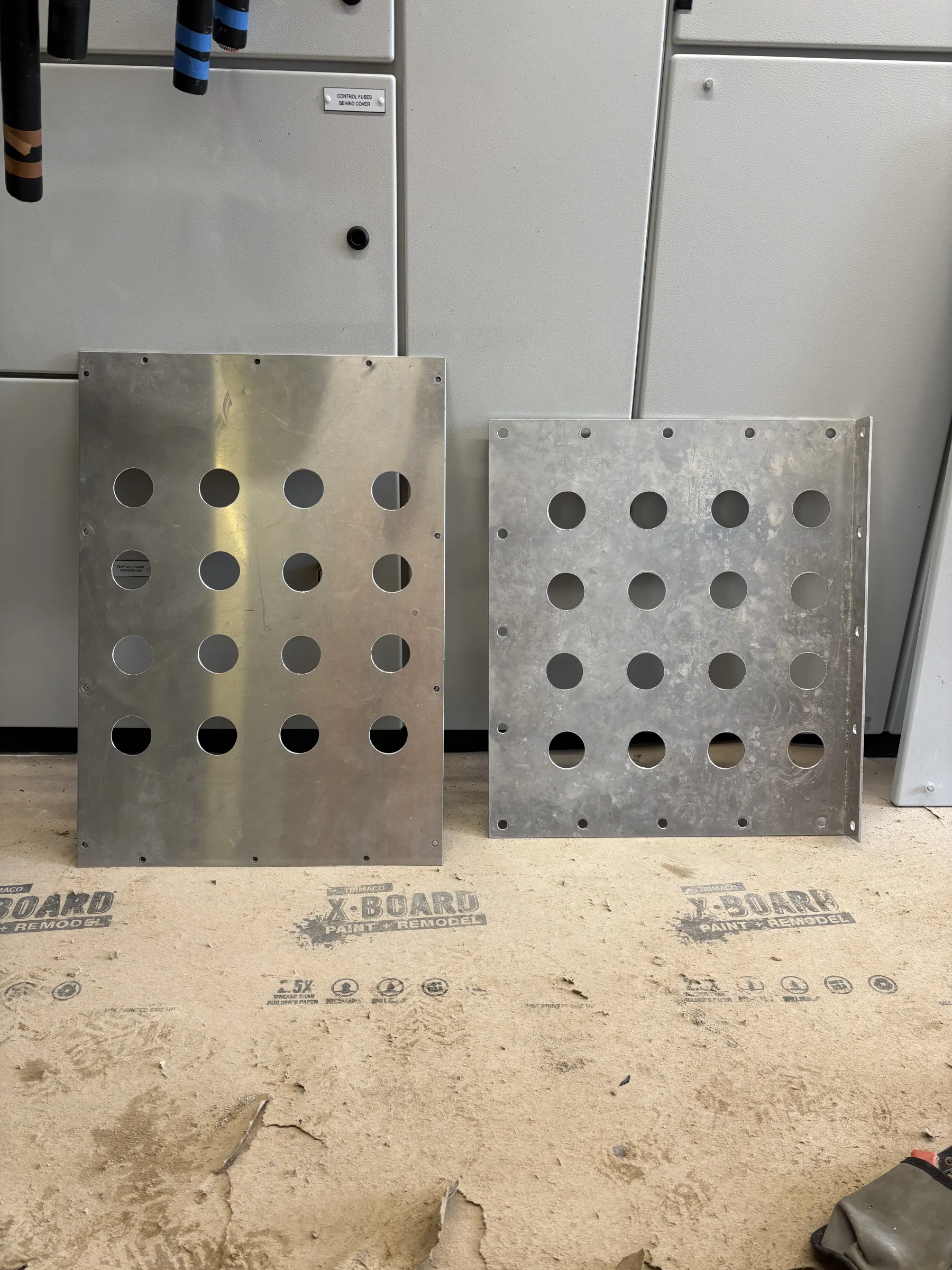 Two rectangular metal plates with rows of circular cut-outs leaning against grey electrical cabinets.