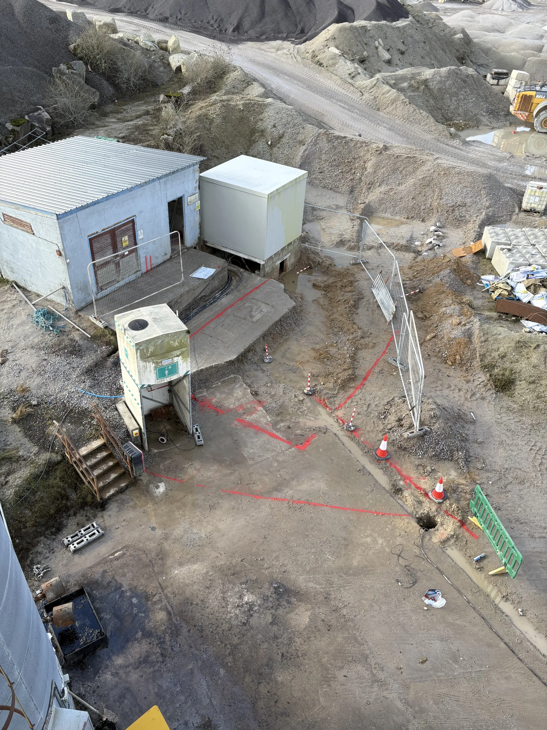 High-angle view of a gravel industrial compound with two utility buildings, a raised cabinet, temporary fencing, cones and red ground markings.