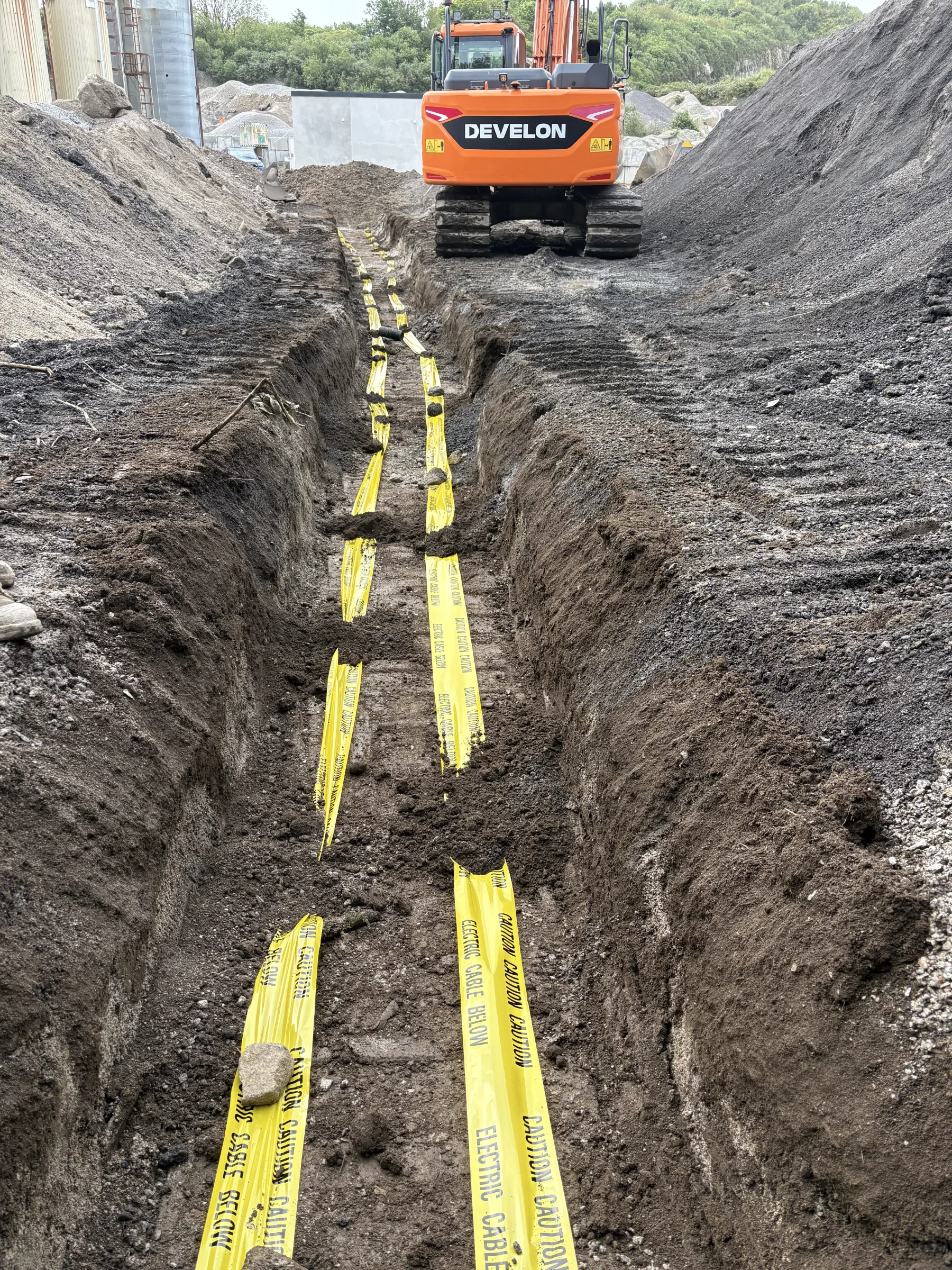 Deep excavated trench containing several lengths of yellow electrical cable warning tape, with a tracked excavator at the far end.