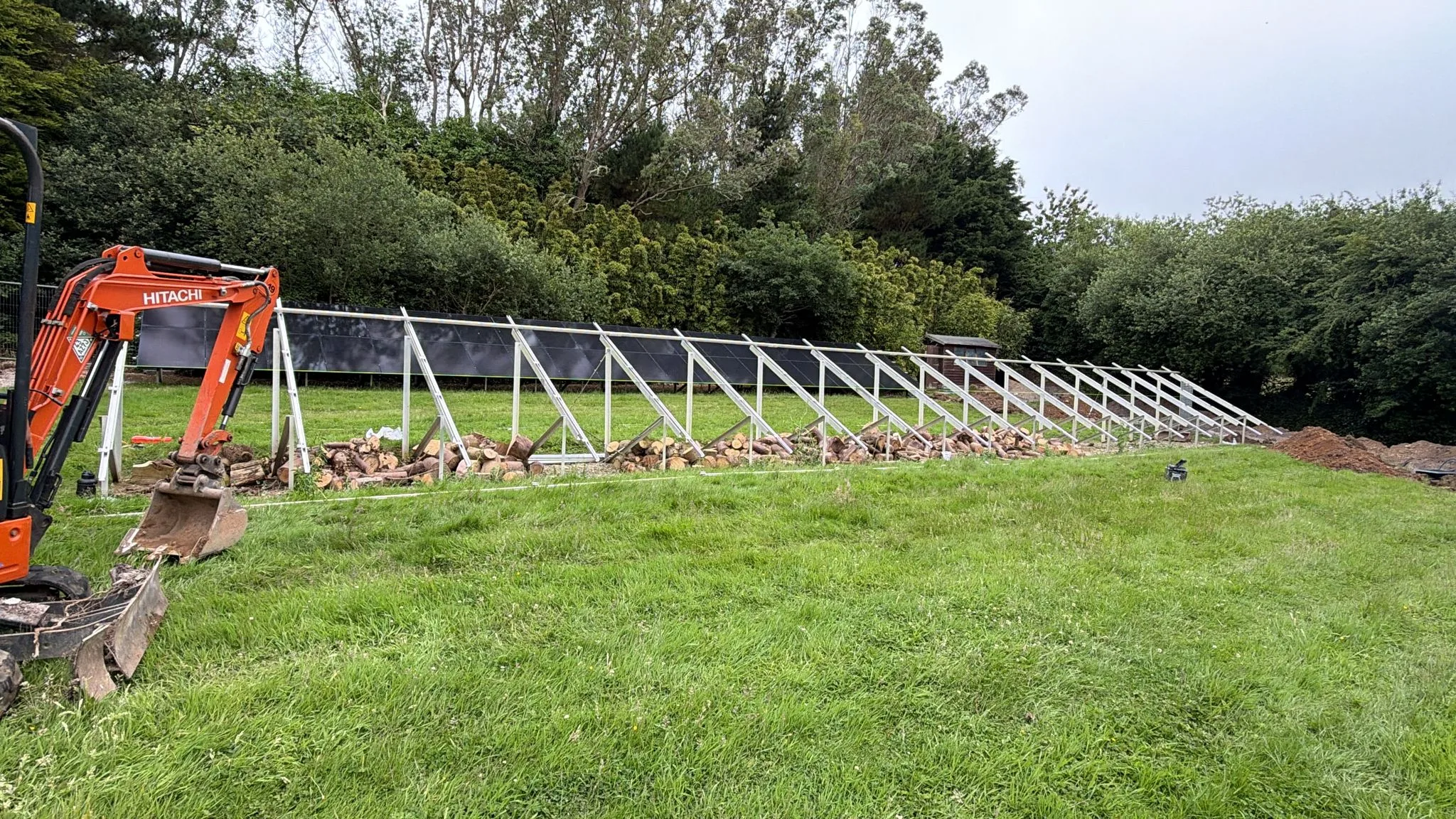 Long row of angled metal solar panel support frames under construction in a grassy field, with an orange mini excavator at the left.