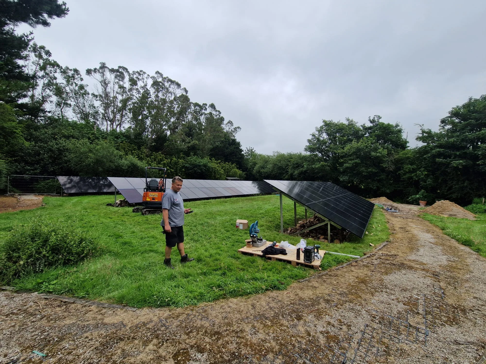 A worker standing on grass beside rows of ground-mounted solar panels, tools, a small orange excavator and a gravel track.