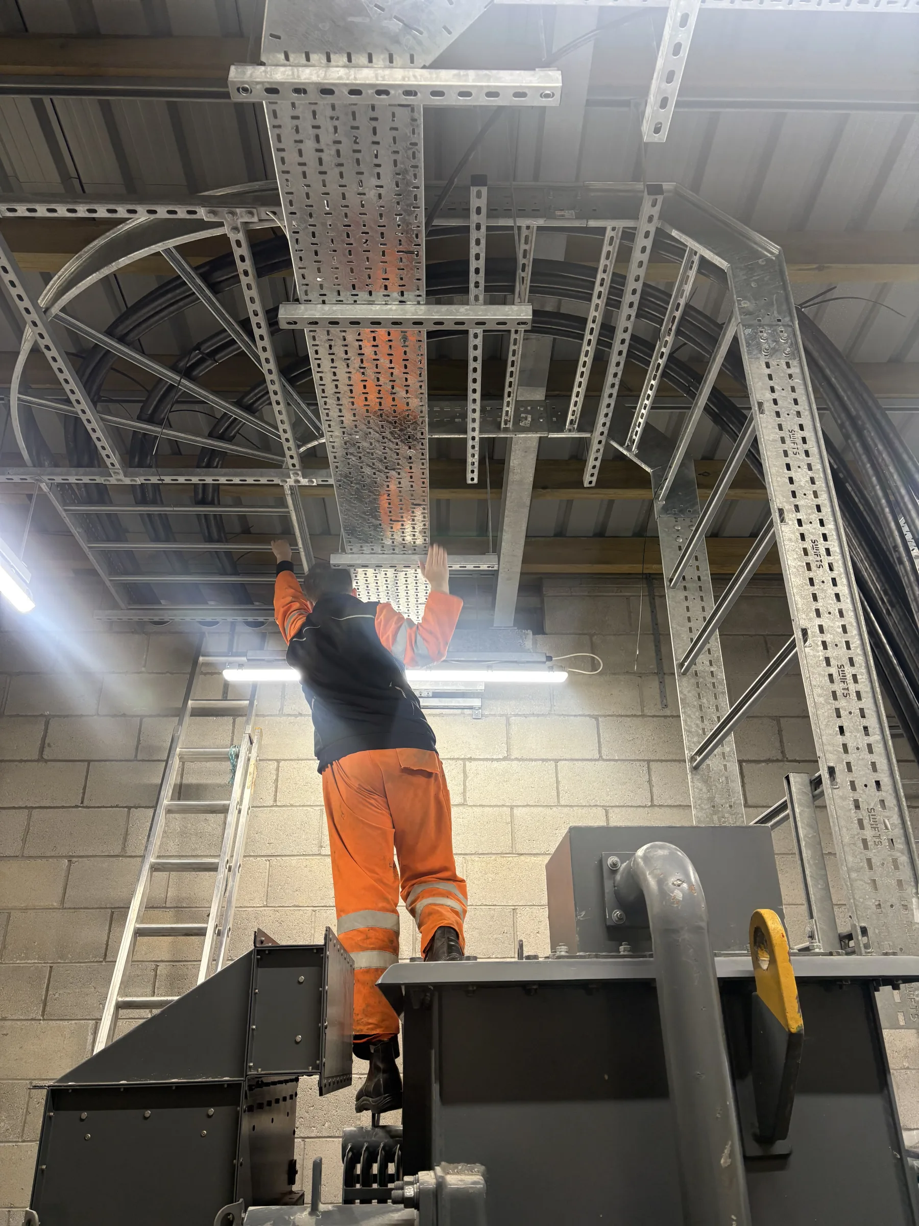 Worker in orange high-visibility trousers fitting perforated metal cable tray to an overhead support system inside an industrial building.