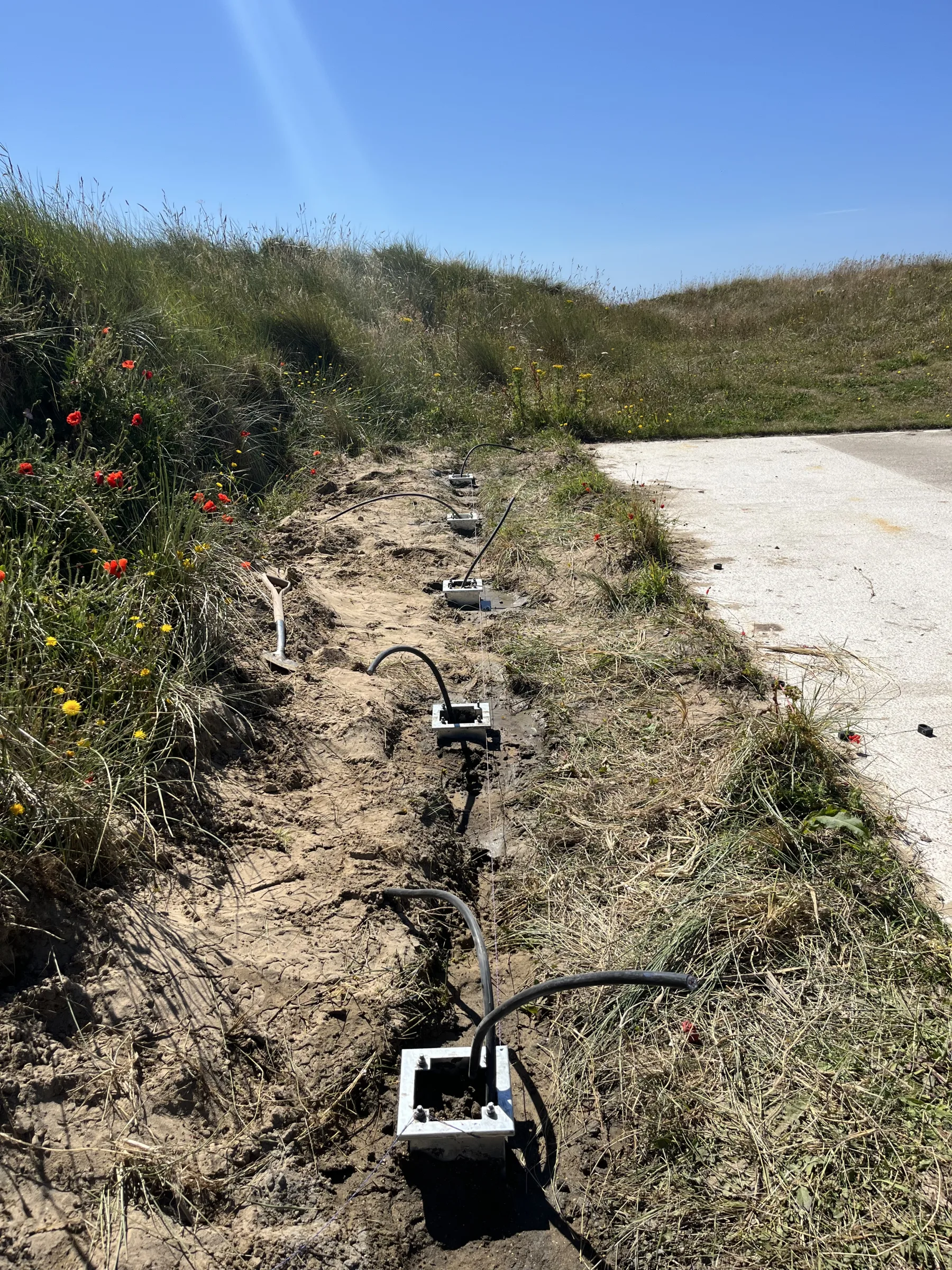 An open trench beside a paved area contains several square post bases and loops of black cable, with grass and wildflowers alongside.