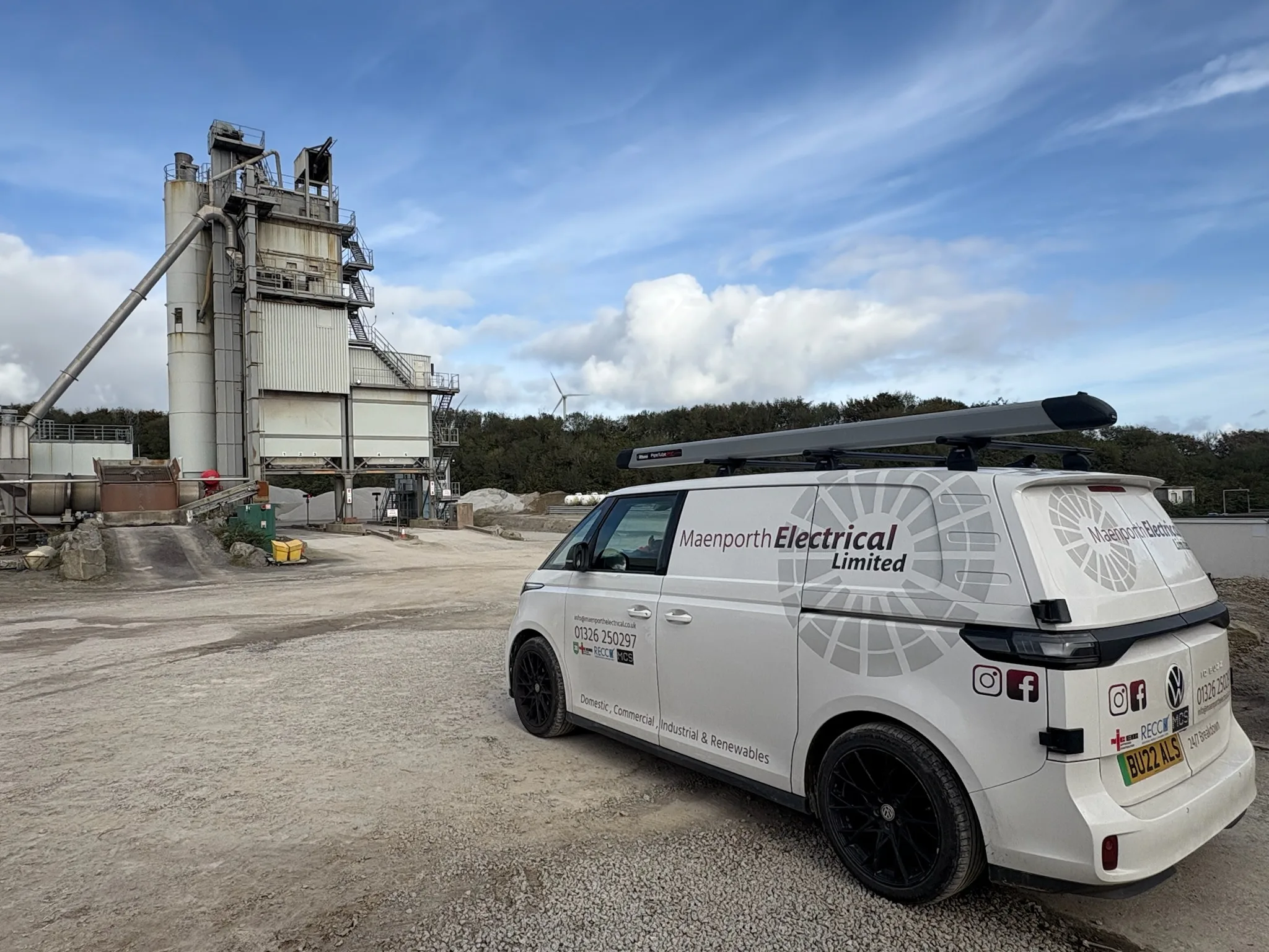 Rear three-quarter view of a white Maenporth Electrical van with roof tubes, parked near tall industrial processing equipment.