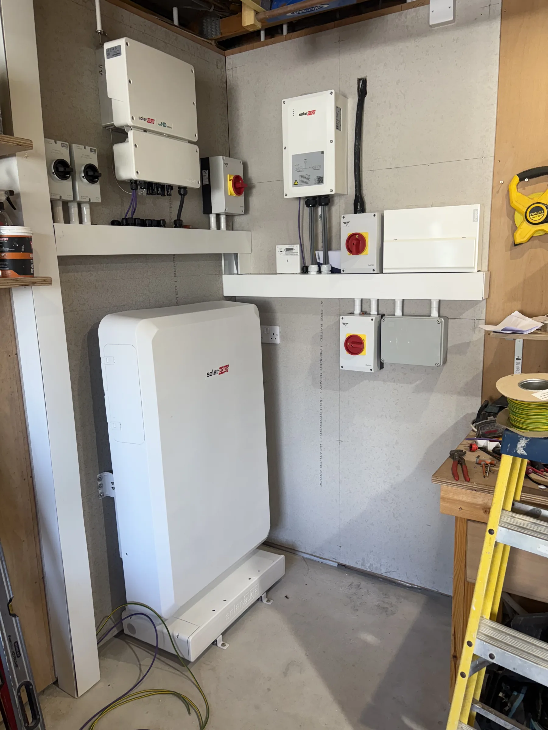 White battery and inverter units mounted against a utility room wall with conduit, electrical enclosures and red rotary isolators.