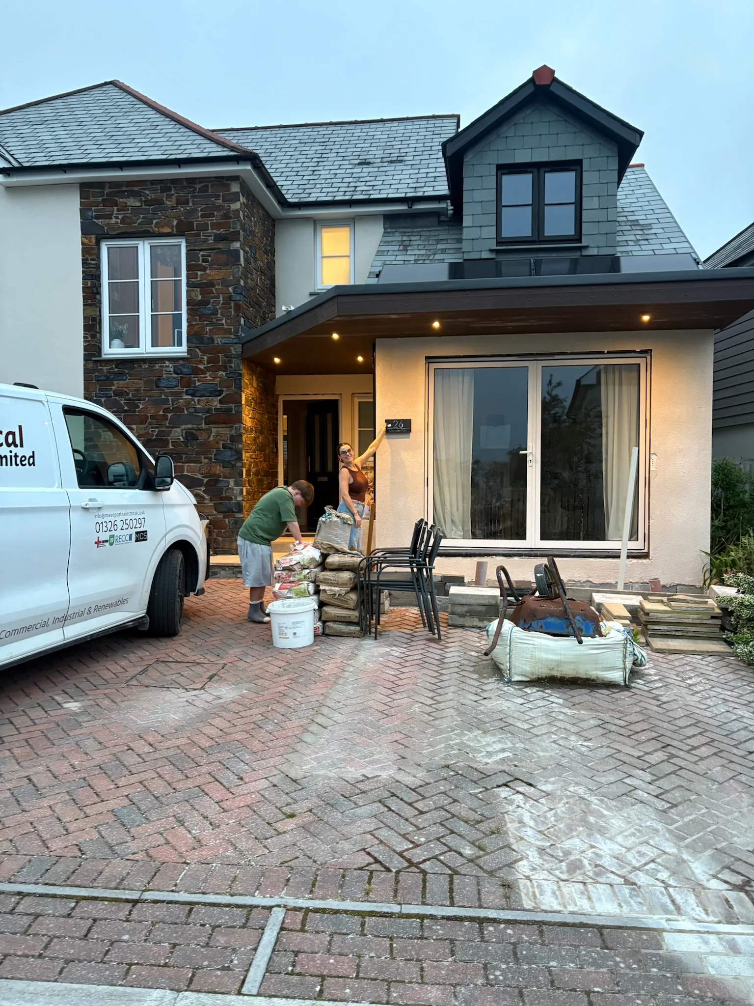 House with illuminated recessed exterior lights, a white work van, two people and building materials arranged on a paved driveway.
