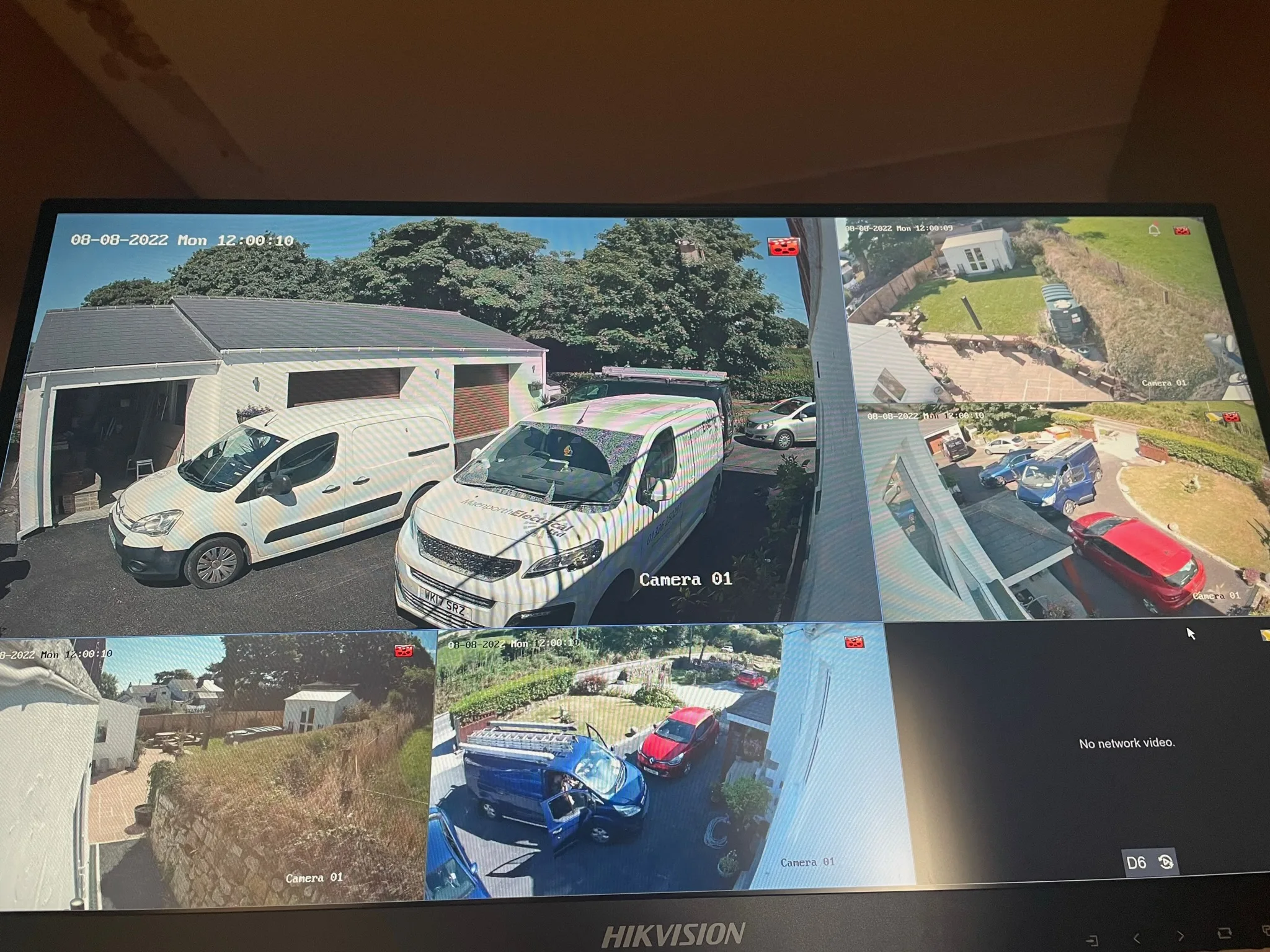 Monitor showing a multi-camera CCTV layout with views of a house, driveway, garden, work vans and parked cars, plus one inactive camera panel.
