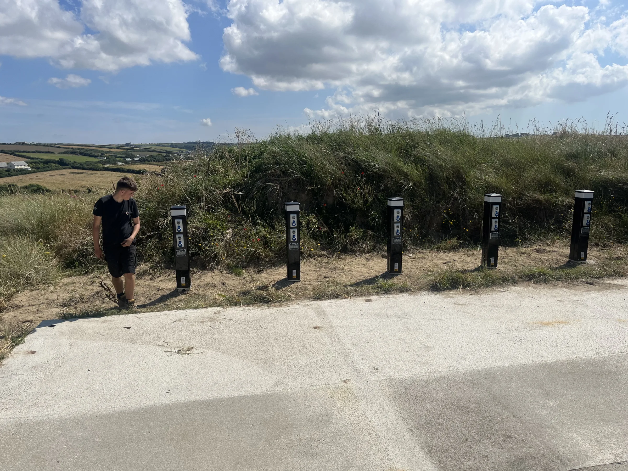 A person stands beside five black electrical posts with pale tops and front-mounted sockets, arranged along a grassy verge beside paving.