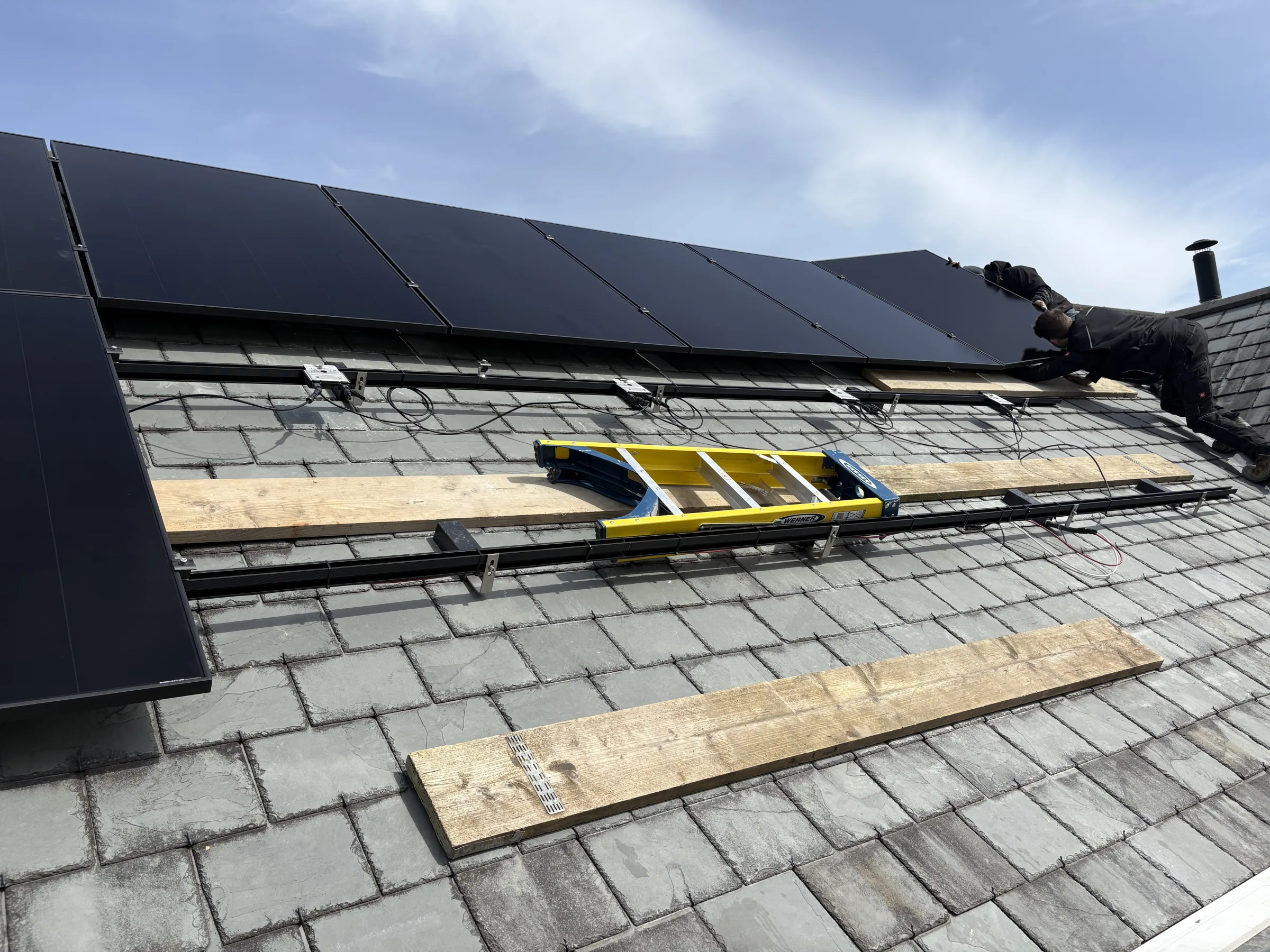 Rows of dark solar panels above exposed mounting rails and cables on a slate roof, with a yellow ladder and timber boards laid across the tiles.