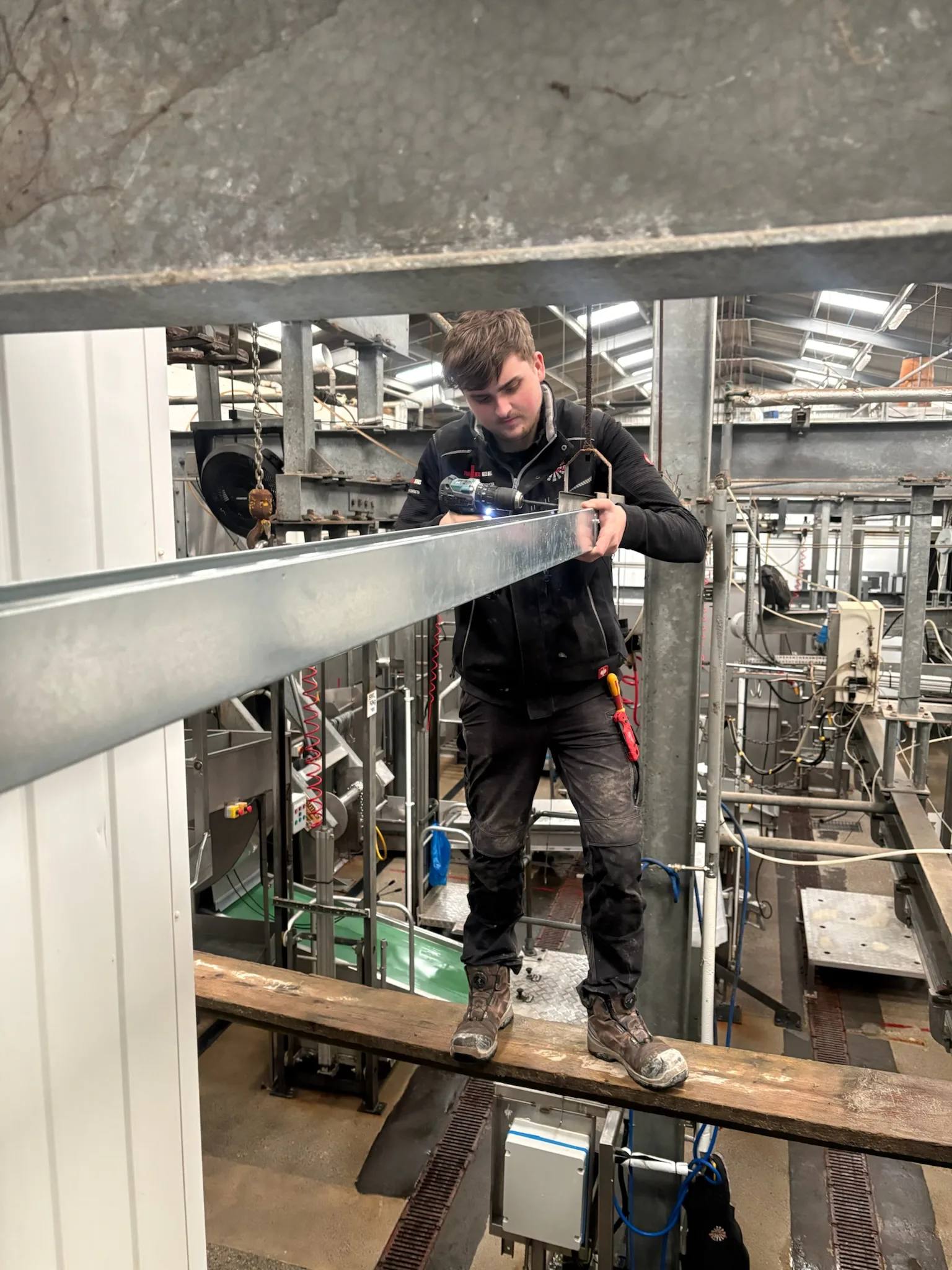 Electrician standing on a timber board at height and using a hand tool on a long galvanised metal channel in an industrial interior.
