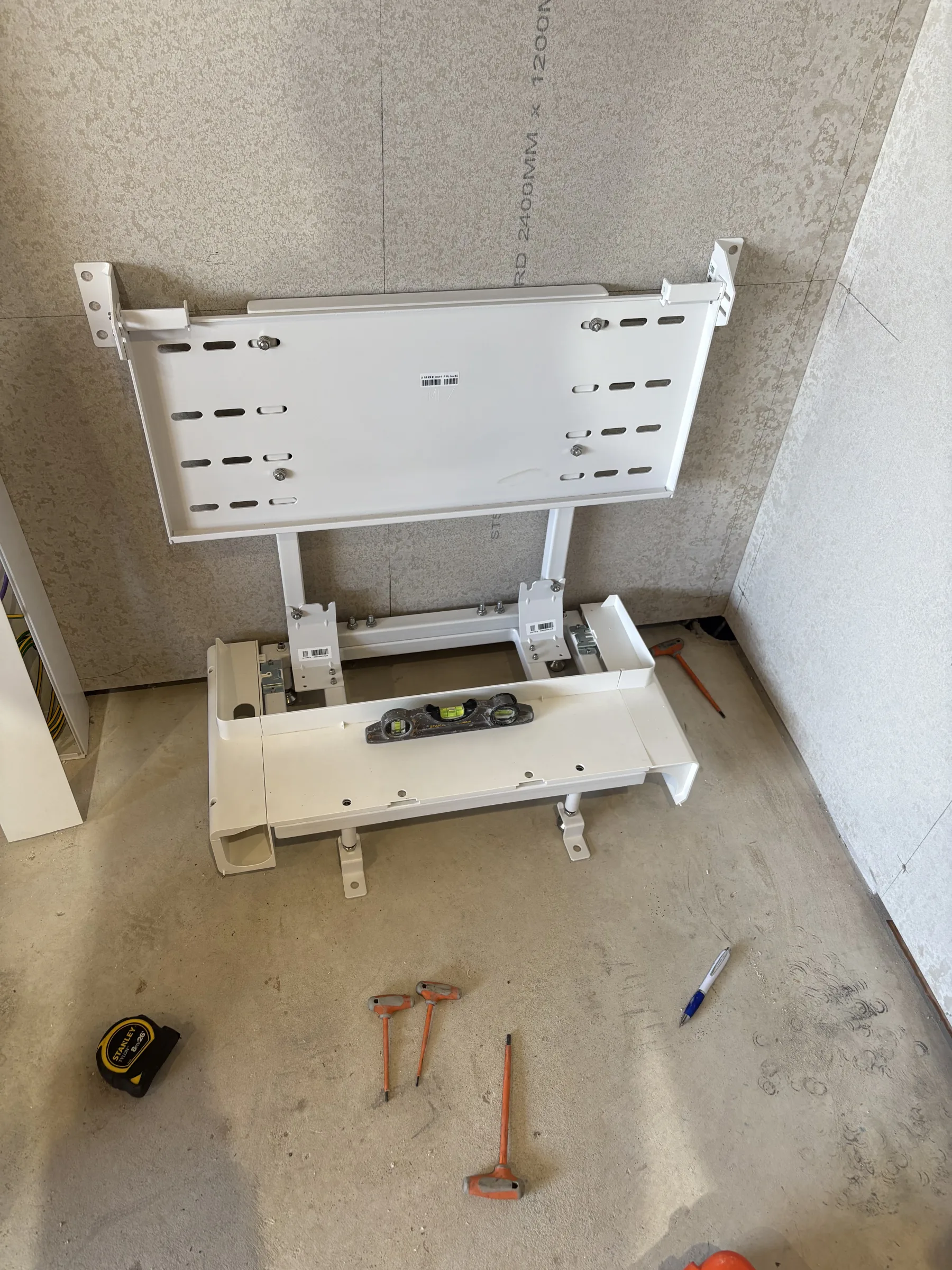 Two white metal equipment mounting brackets fixed to an unfinished block wall, with hand tools on the concrete floor below.