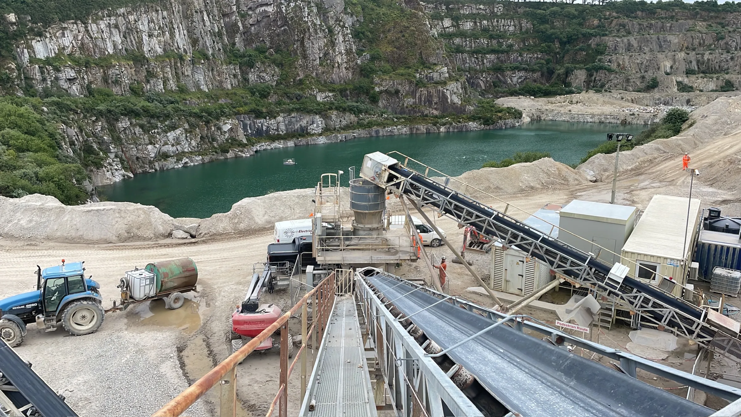 Elevated view along metal conveyor belts across a quarry processing site, with a tractor and water-filled quarry pit below.