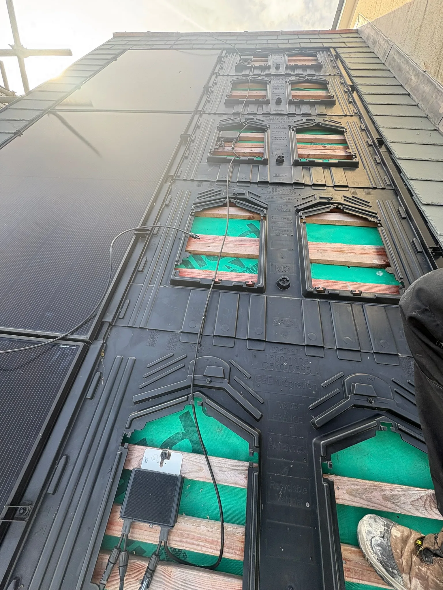 View up a pitched roof showing black mounting trays, open sections revealing green membrane and timber battens, loose cables and the edge of a dark panel.
