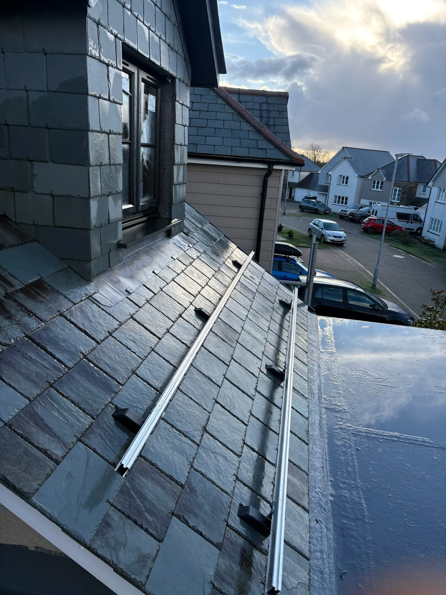 Parallel metal rails and black brackets on a pitched slate roof beneath a slate-clad dormer, overlooking a residential street.