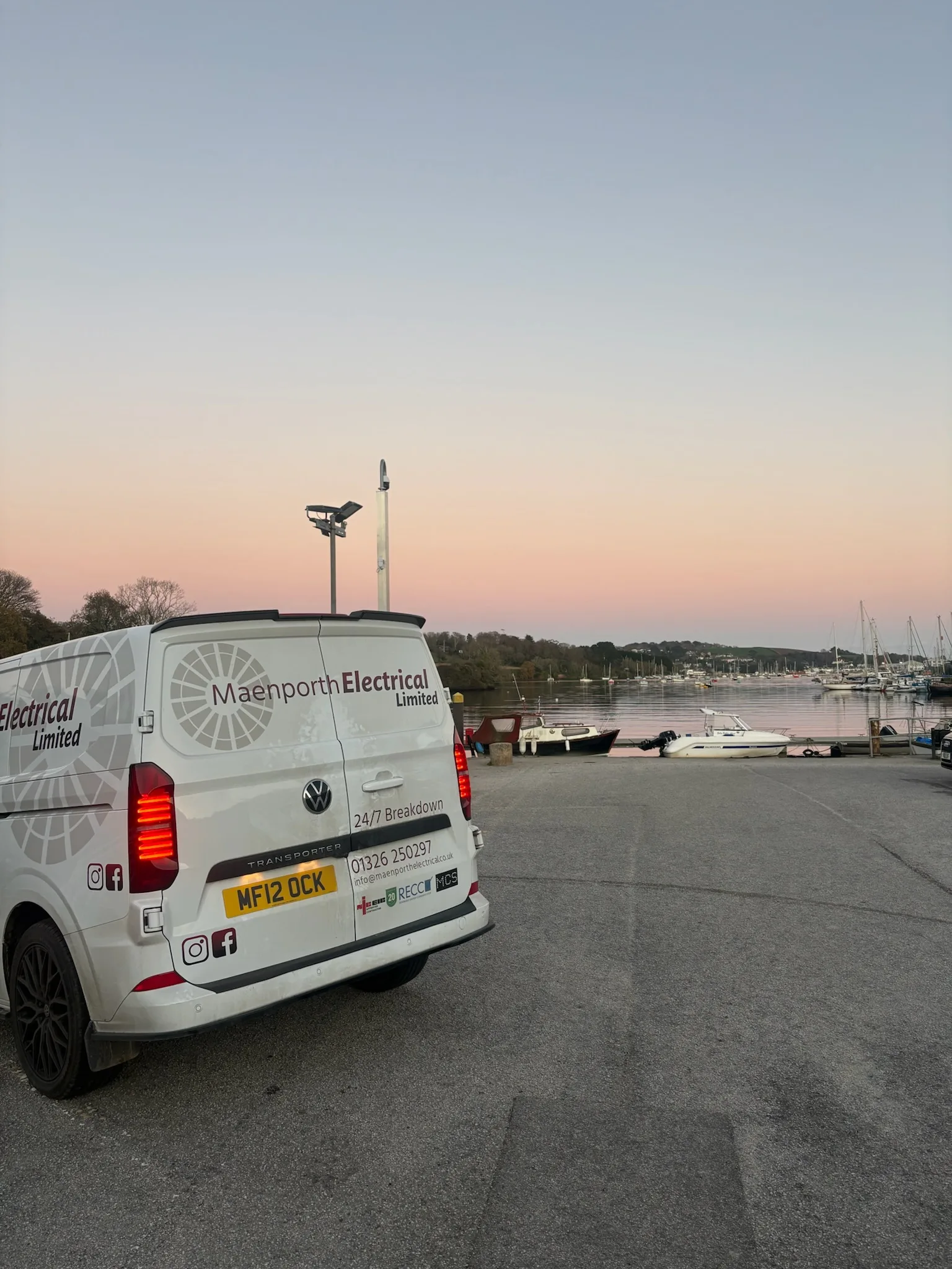 Rear of a white Maenporth Electrical van parked above a harbour containing small boats and sailing masts at sunset.