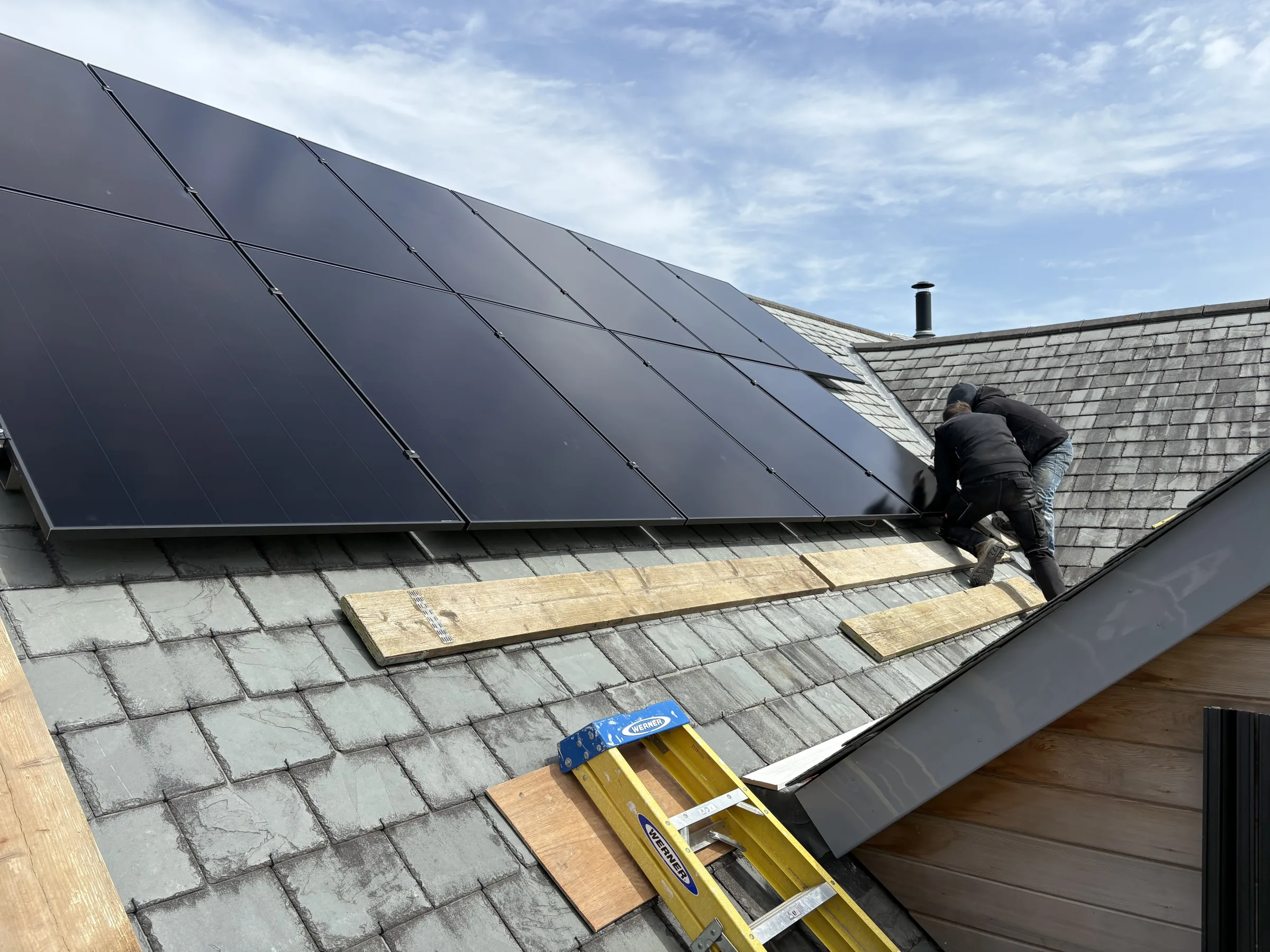 Dark solar panels fitted across a slate roof, with an installer working beside the array and a yellow ladder below.