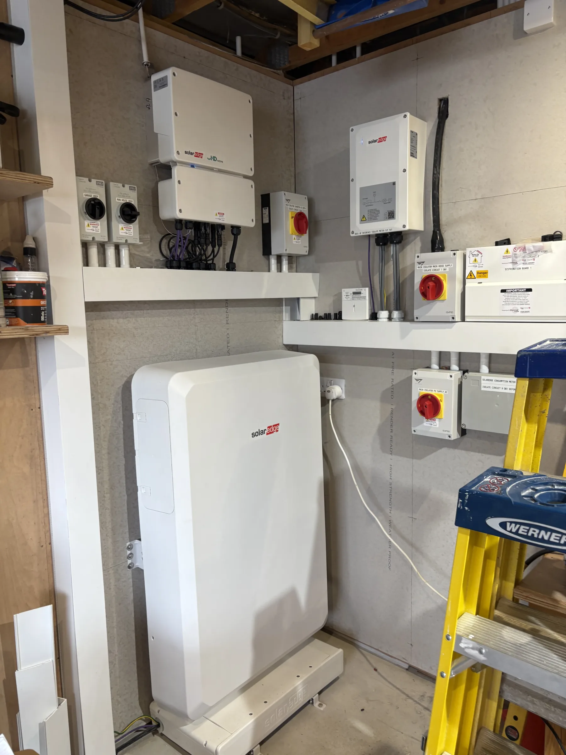 Floor-standing white battery unit below wall-mounted electrical equipment, isolator switches and cable trunking, with a yellow step ladder at the right.