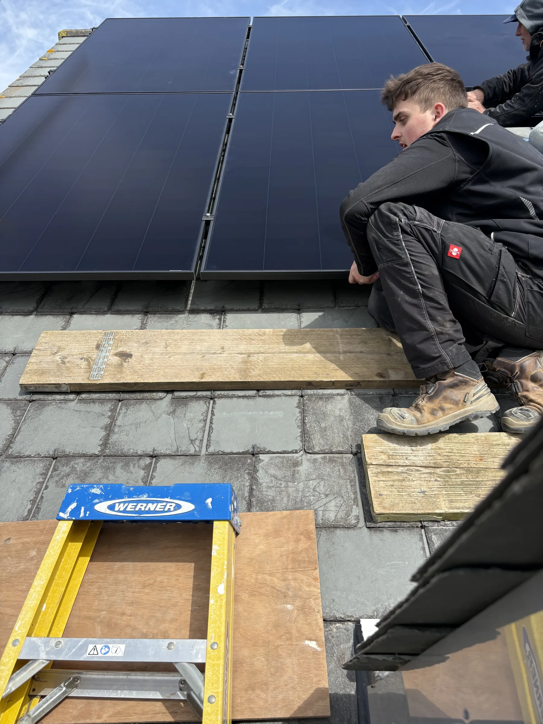 Installer crouching on a slate roof while holding the lower edge of a dark solar panel, with another worker partly visible behind.