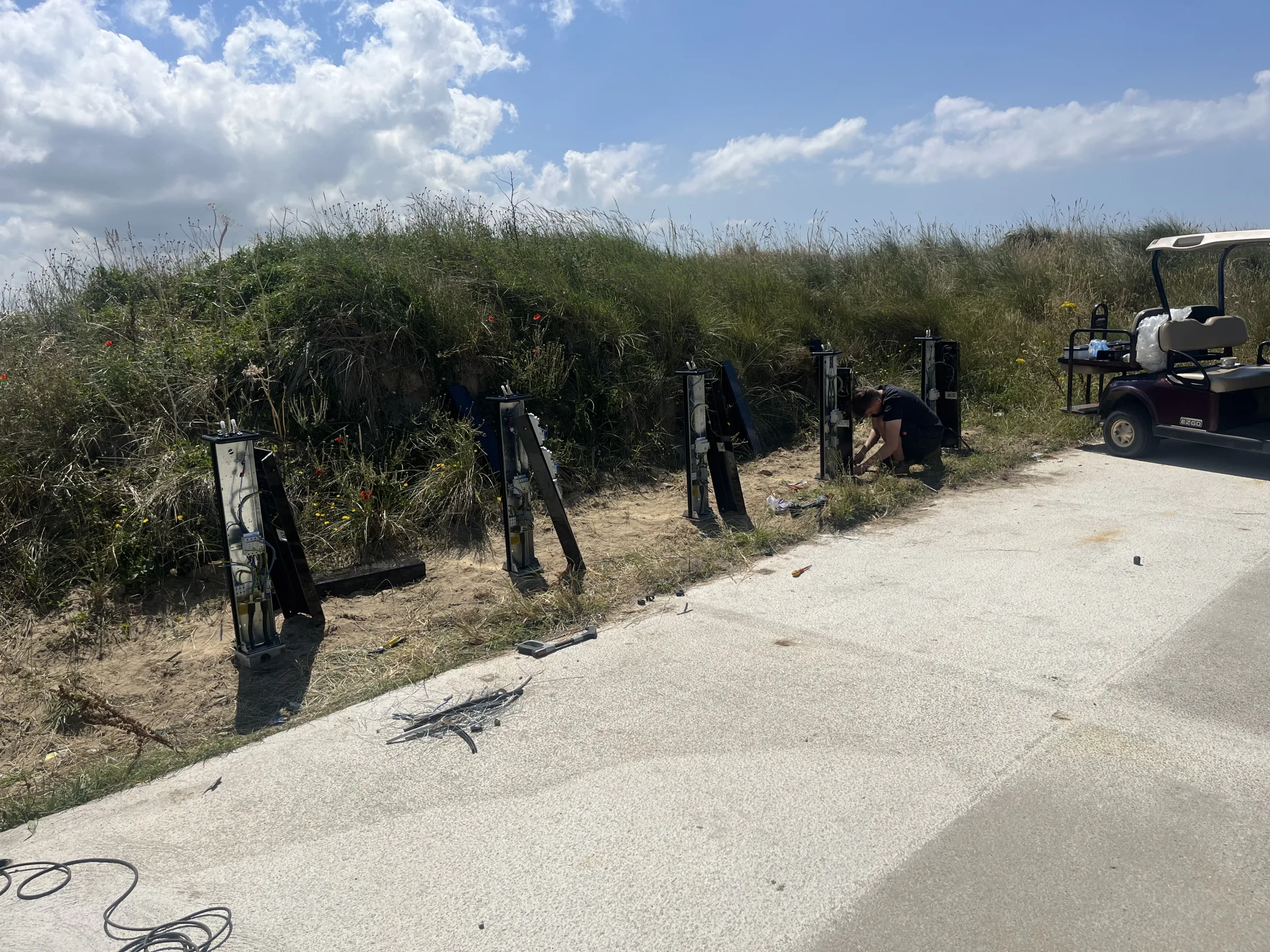 Several upright black housings with exposed internal sections stand along a grassy bank beside a surfaced path. A person crouches near one housing, with a utility buggy to the right.