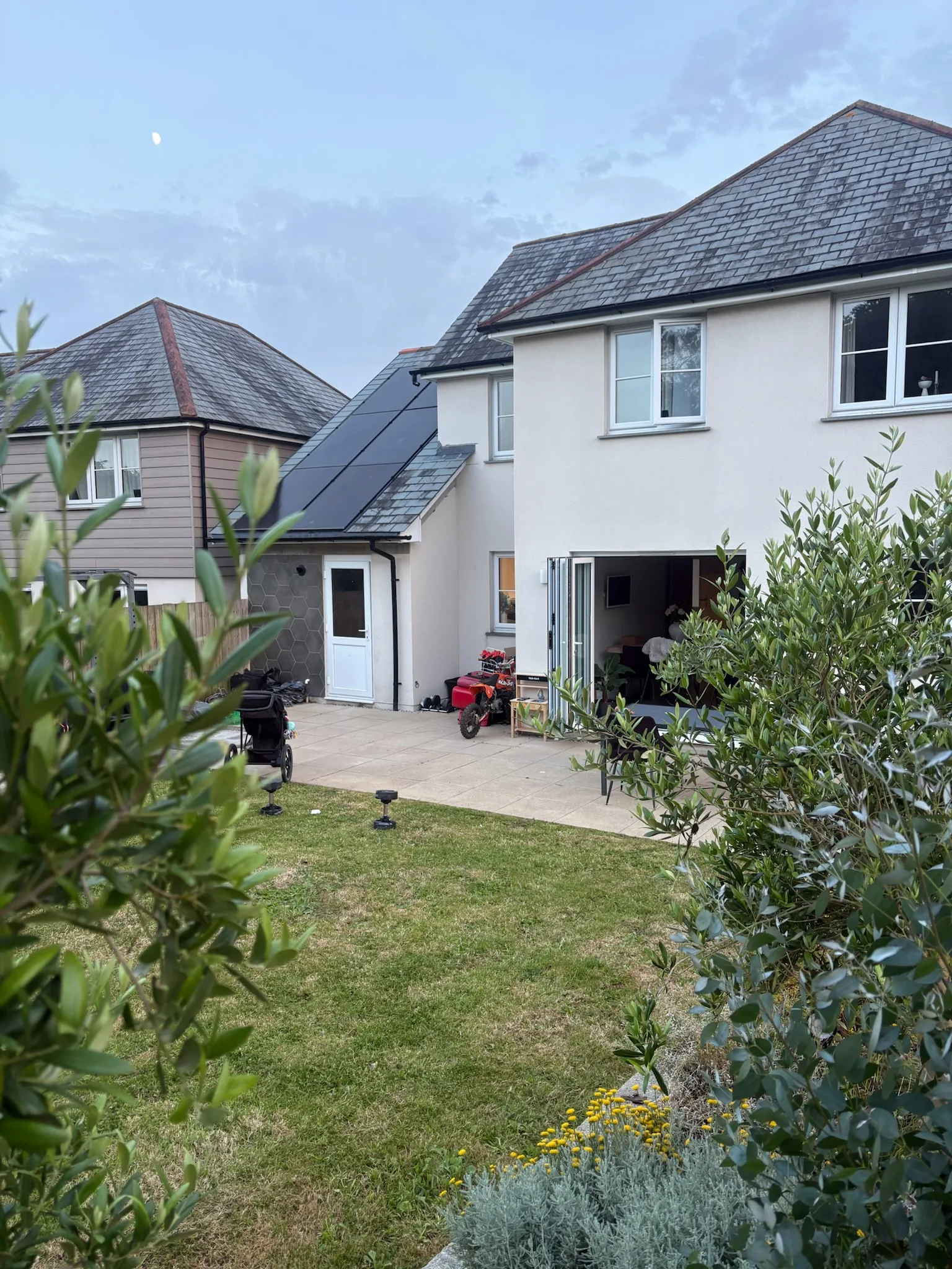 Rear and side of a two-storey house seen through garden shrubs, with dark solar panels on the left-hand pitched roof.