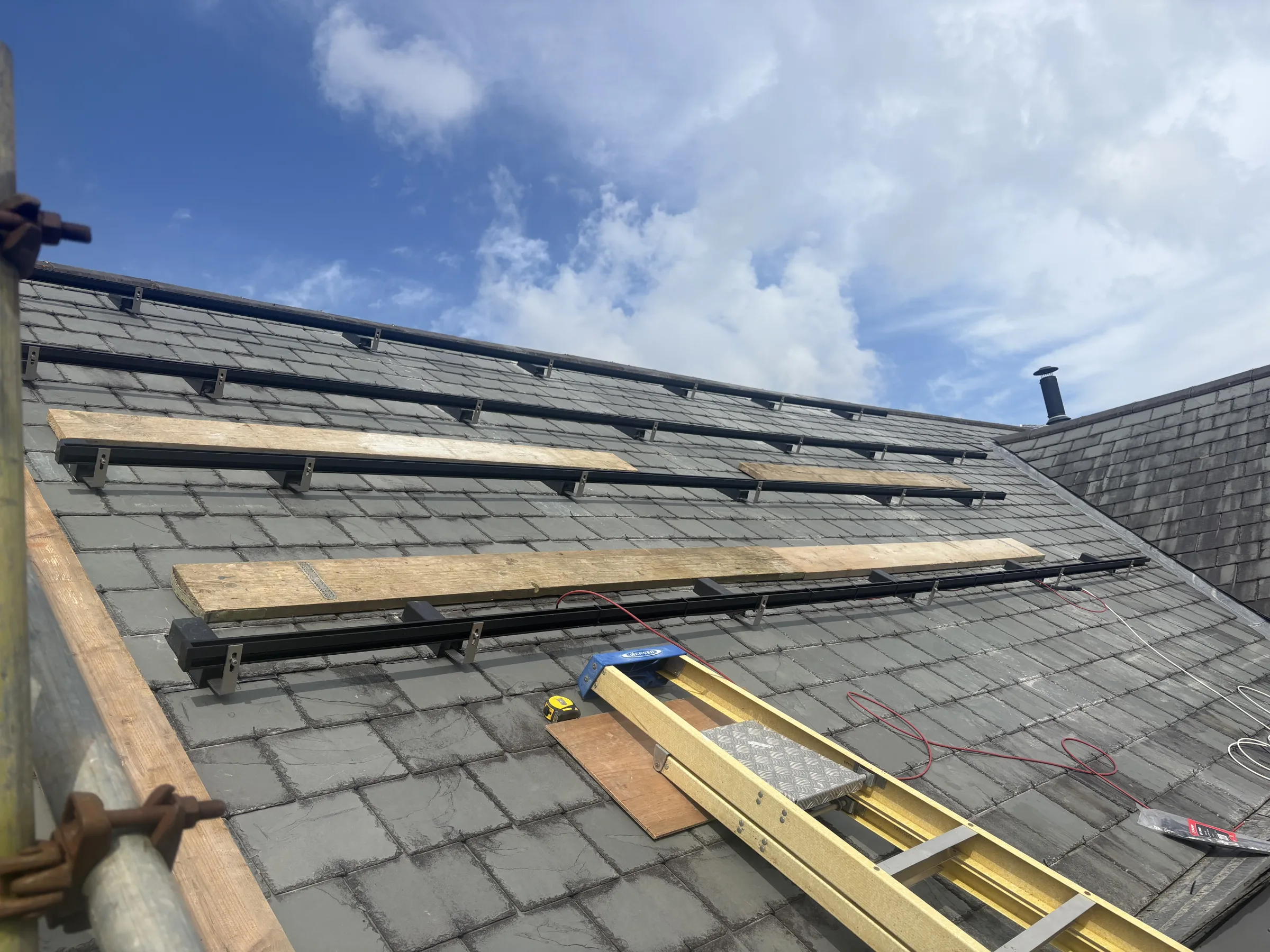 Horizontal black mounting rails fixed across a slate roof, with timber boards, a yellow ladder and loose red and black cables visible.