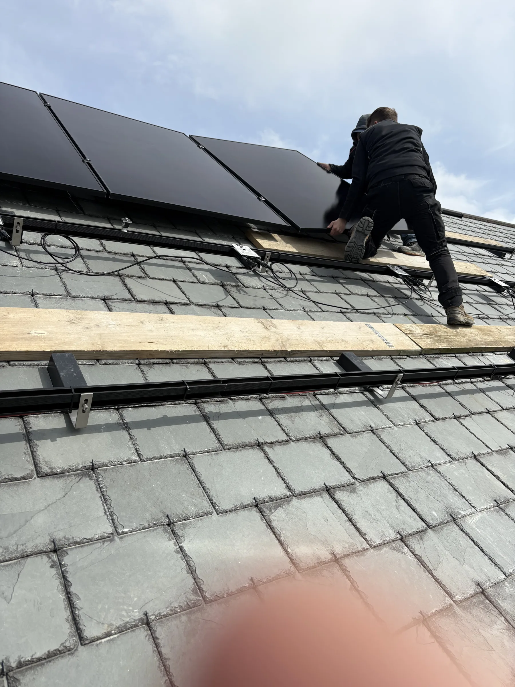 Installer kneeling on a slate roof and holding a dark solar panel above visible mounting rails, cables and timber access boards.