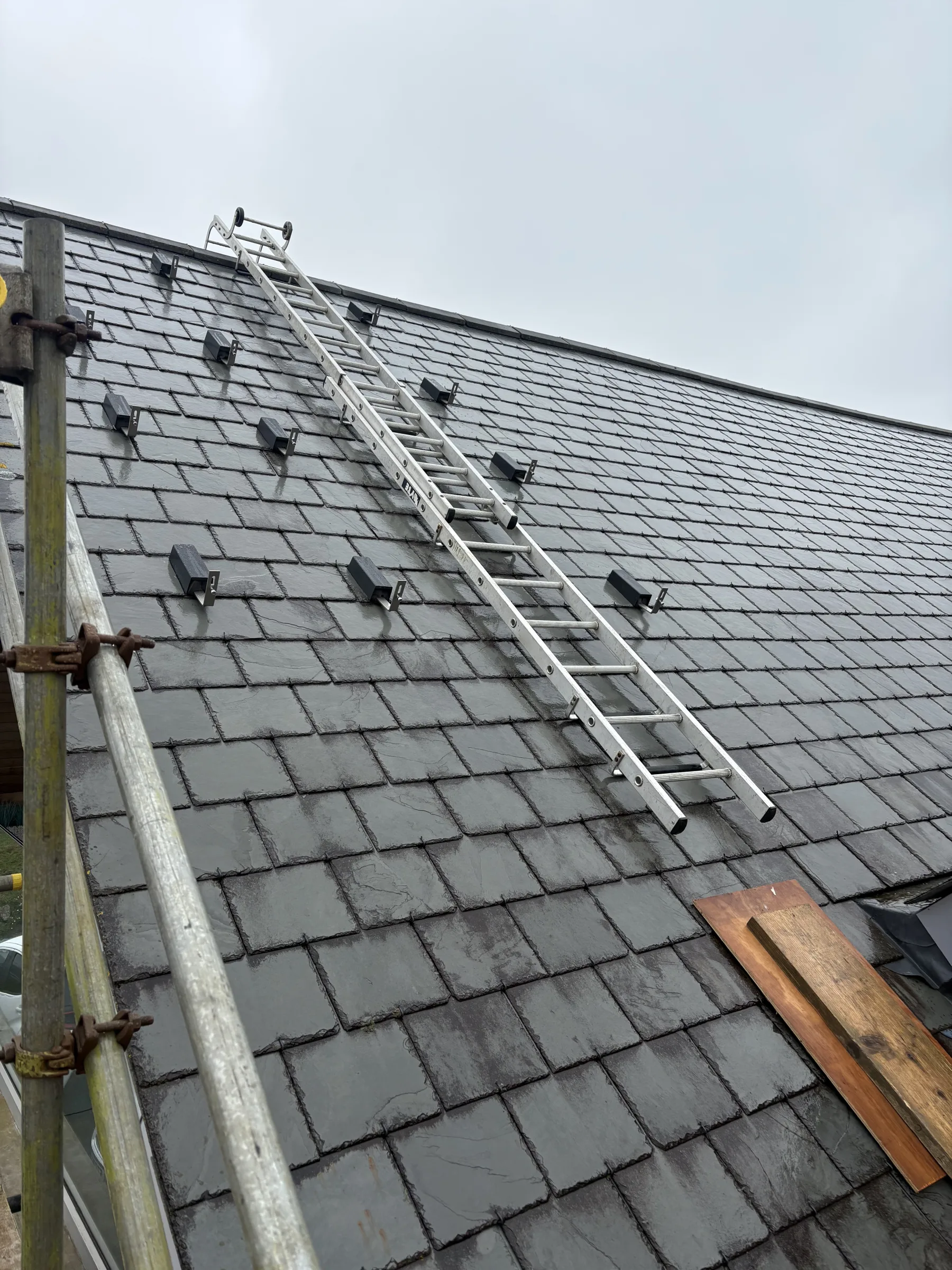 Long aluminium ladder resting on a pitched slate roof fitted with rows of black mounting brackets, with scaffolding at the left.