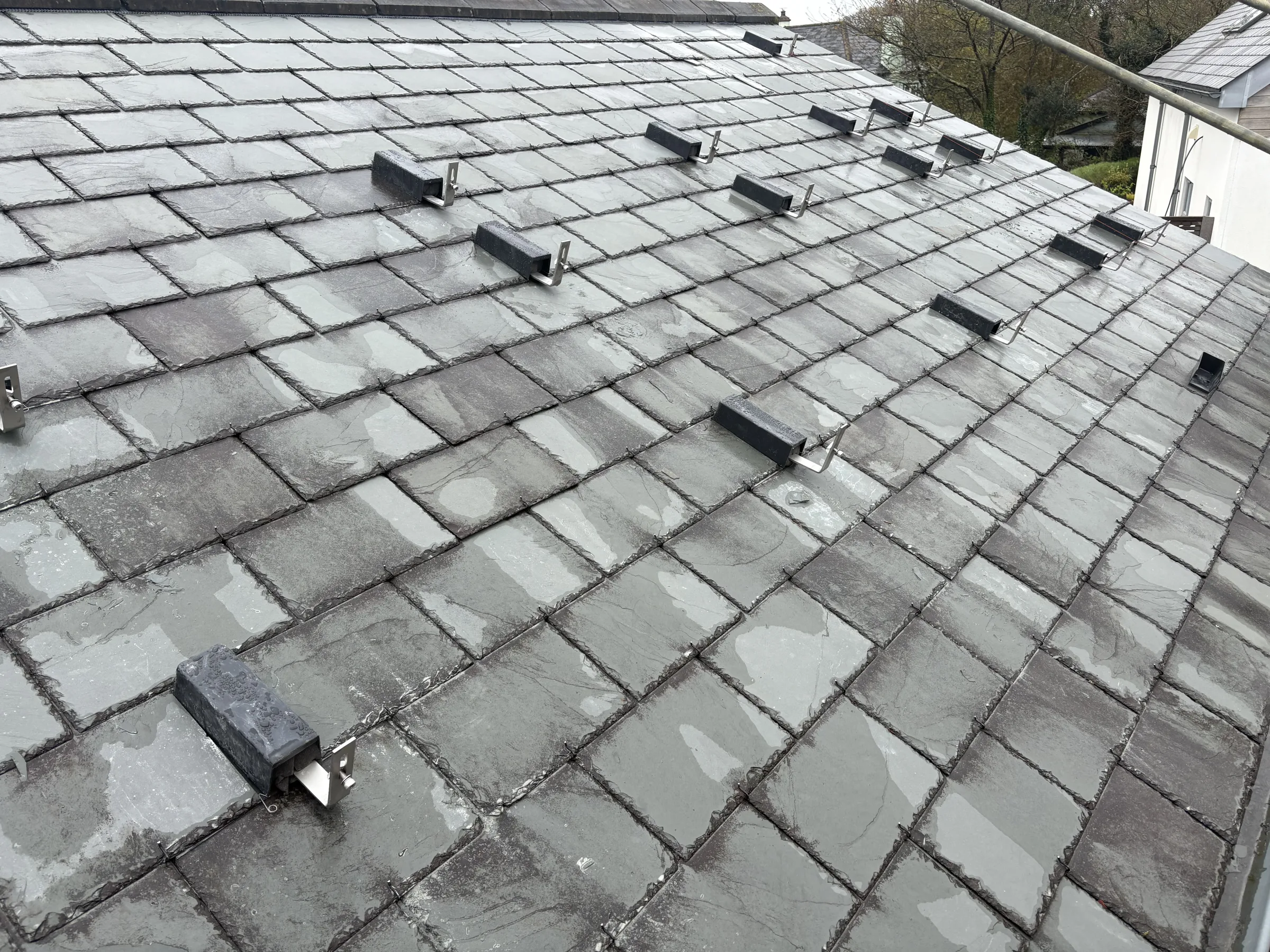 Rows of black metal mounting brackets fixed across wet grey roof slates, viewed from scaffolding.