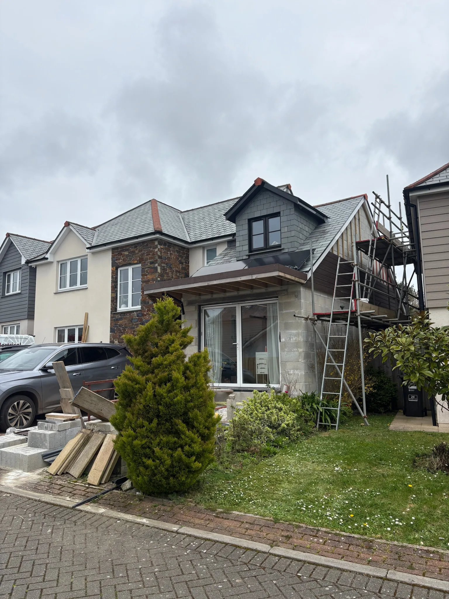 Front of a two-storey house with scaffolding and a ladder at the side, parked cars, stacked building materials and a small lawn.