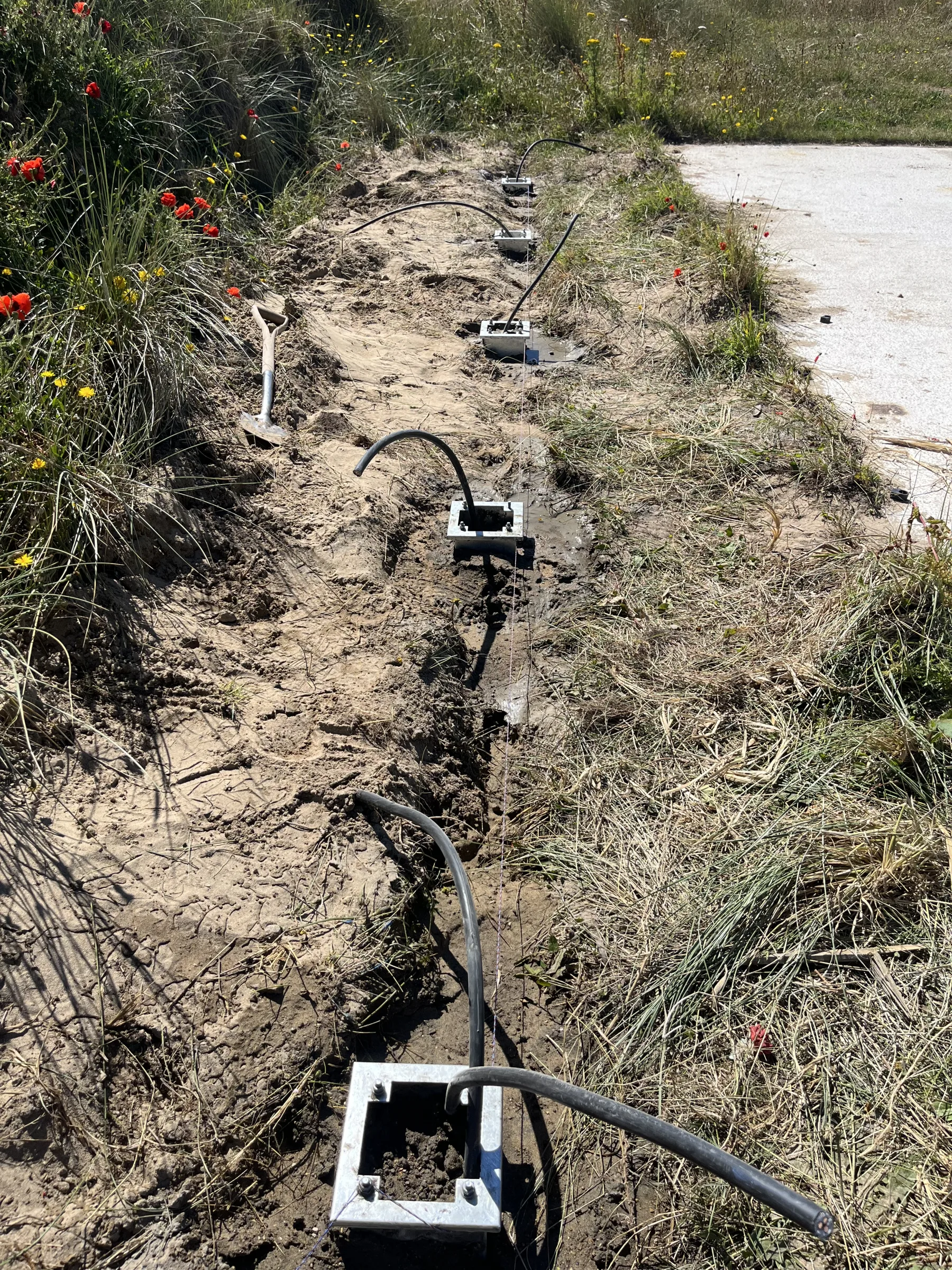 An open trench in sandy soil contains black cables running between square metal mounting bases beside a surfaced path, with a spade resting on the left.