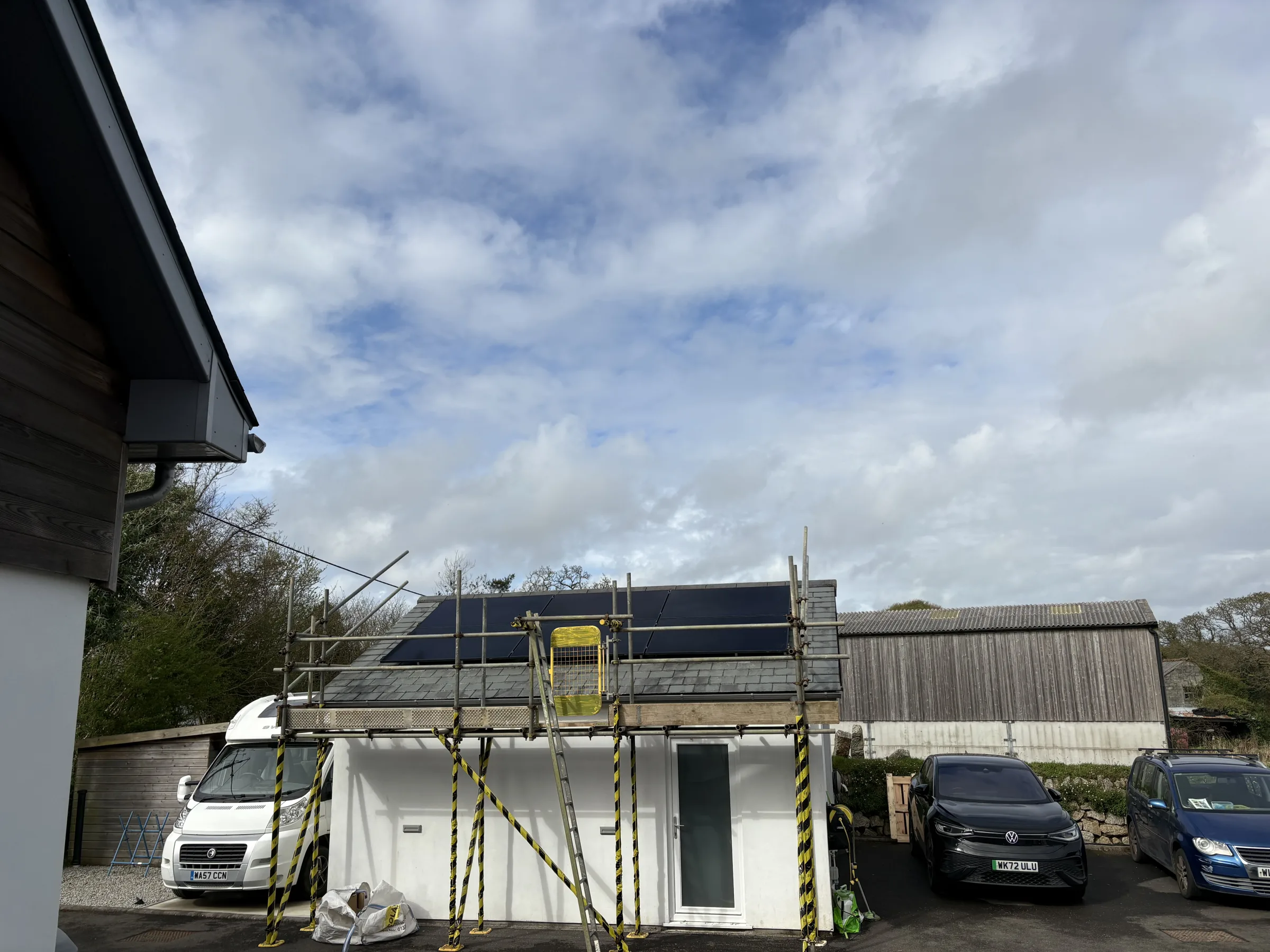 Small slate-roofed building surrounded by scaffolding, with rows of dark solar panels on the roof and vehicles parked nearby.