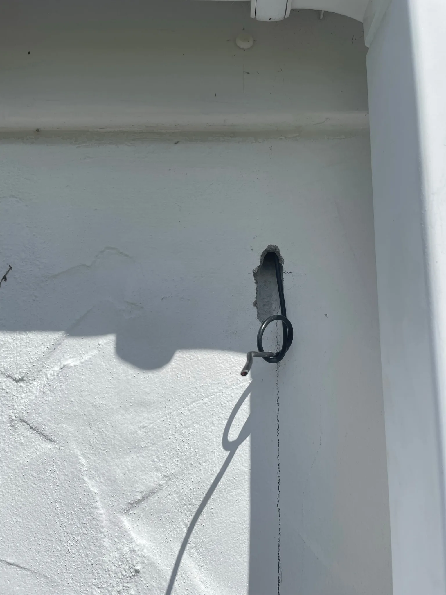 Short loop of dark cable protruding from a narrow opening in a white exterior wall, casting a shadow in bright sunlight.