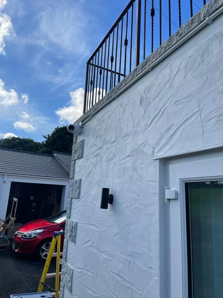 Small cylindrical device mounted high on the corner of a white rendered wall beneath black metal terrace railings, with a wall light below.