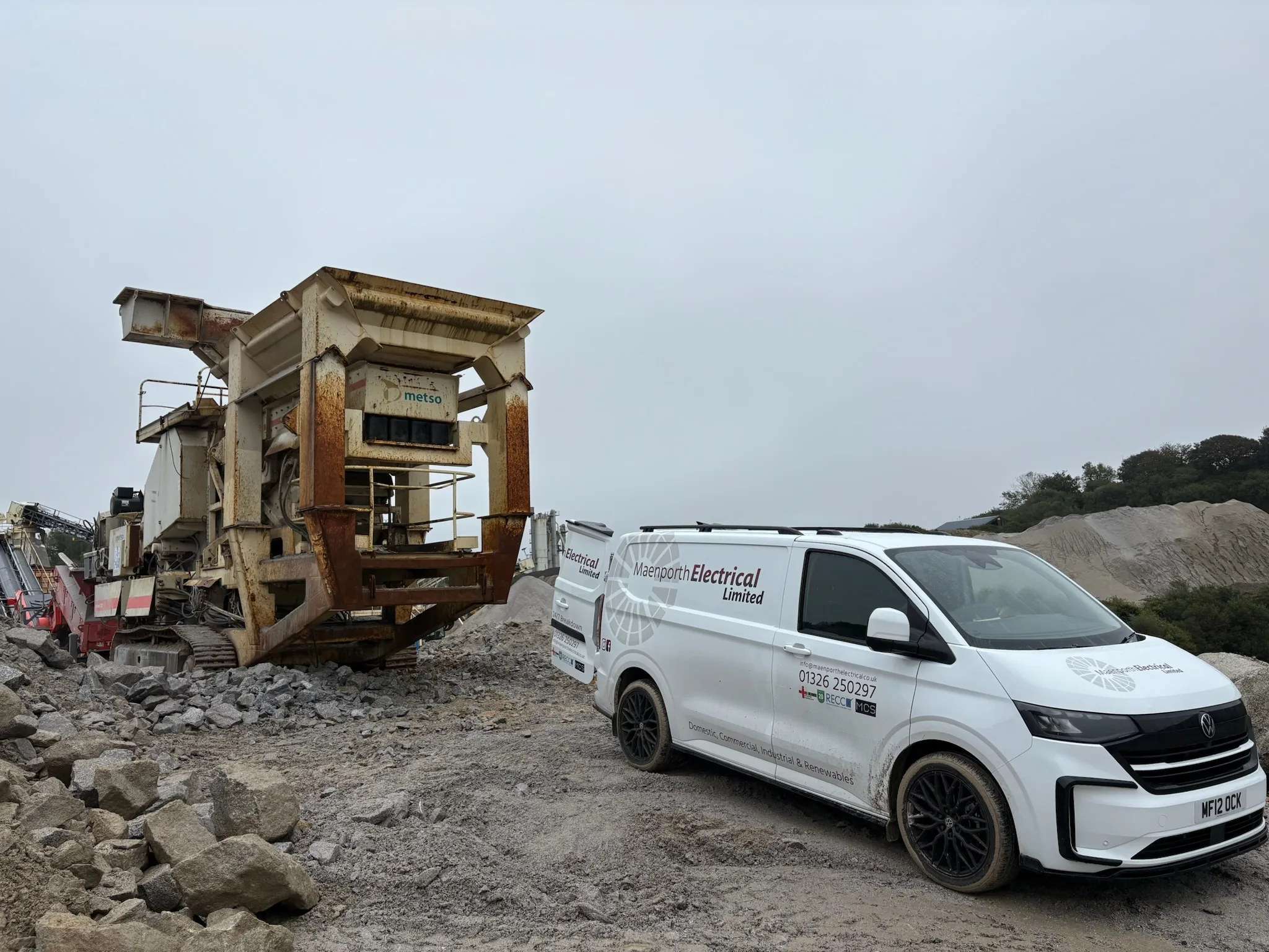 White Maenporth Electrical van parked on rough gravel beside a large tracked crushing machine and piles of stone.
