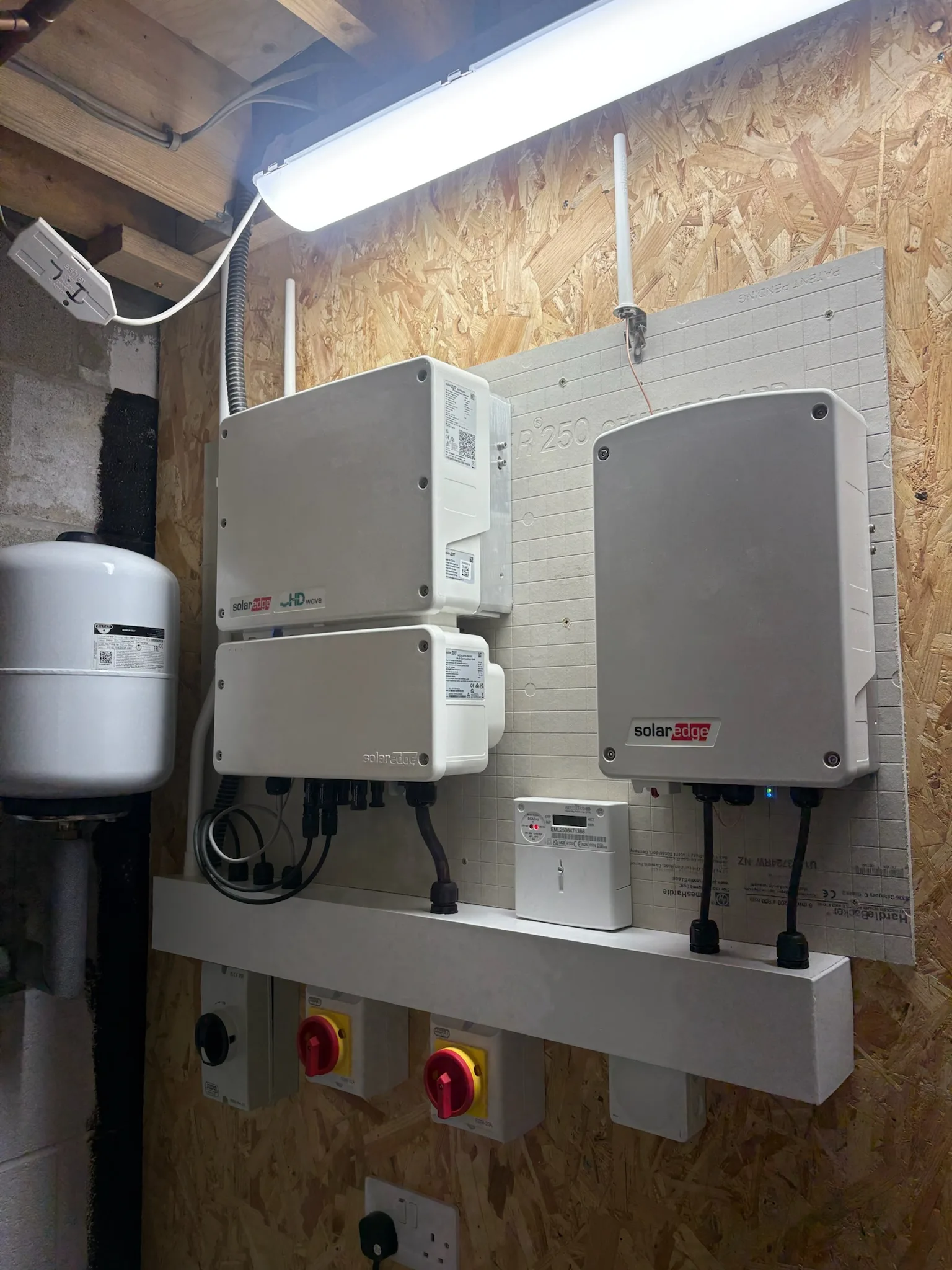 Three grey SolarEdge units with connected cables mounted on a white board in a timber-lined utility space.