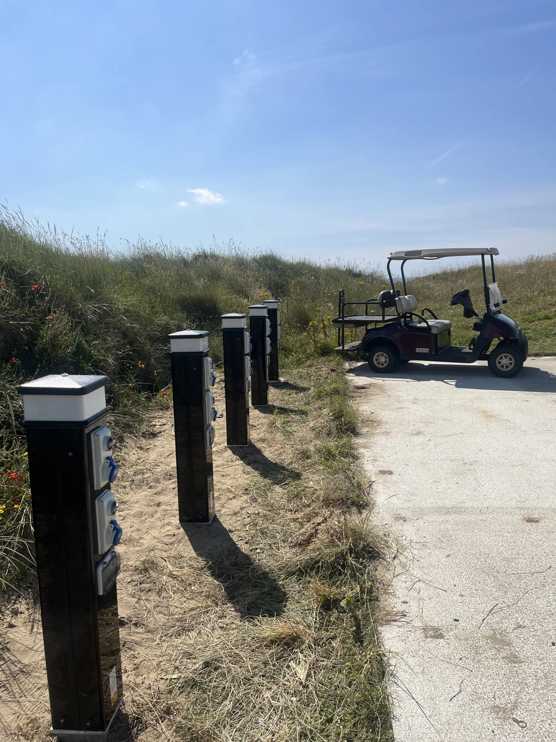 A row of black posts with pale caps and blue covered sockets recedes beside a paved area, with a golf buggy parked at the far end.