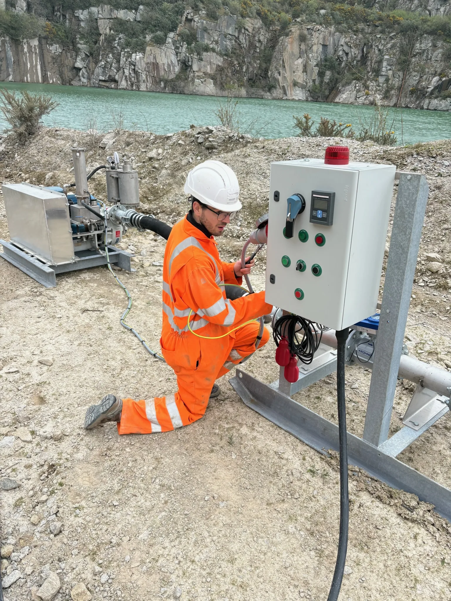Electrician in orange high-visibility clothing and a white hard hat kneeling beside an open electrical control panel in a rocky quarry.
