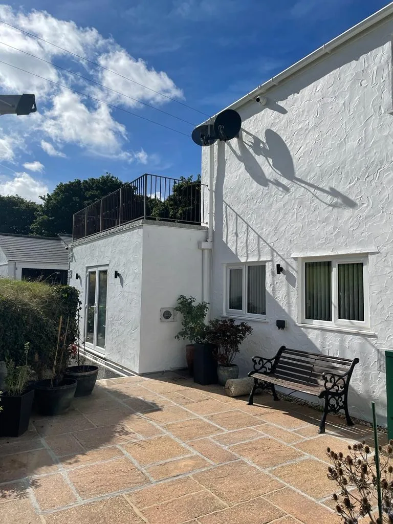 Sunlit paved courtyard beside a white rendered house, with wall-mounted fixtures, two satellite dishes, windows, doors, planters and a bench.
