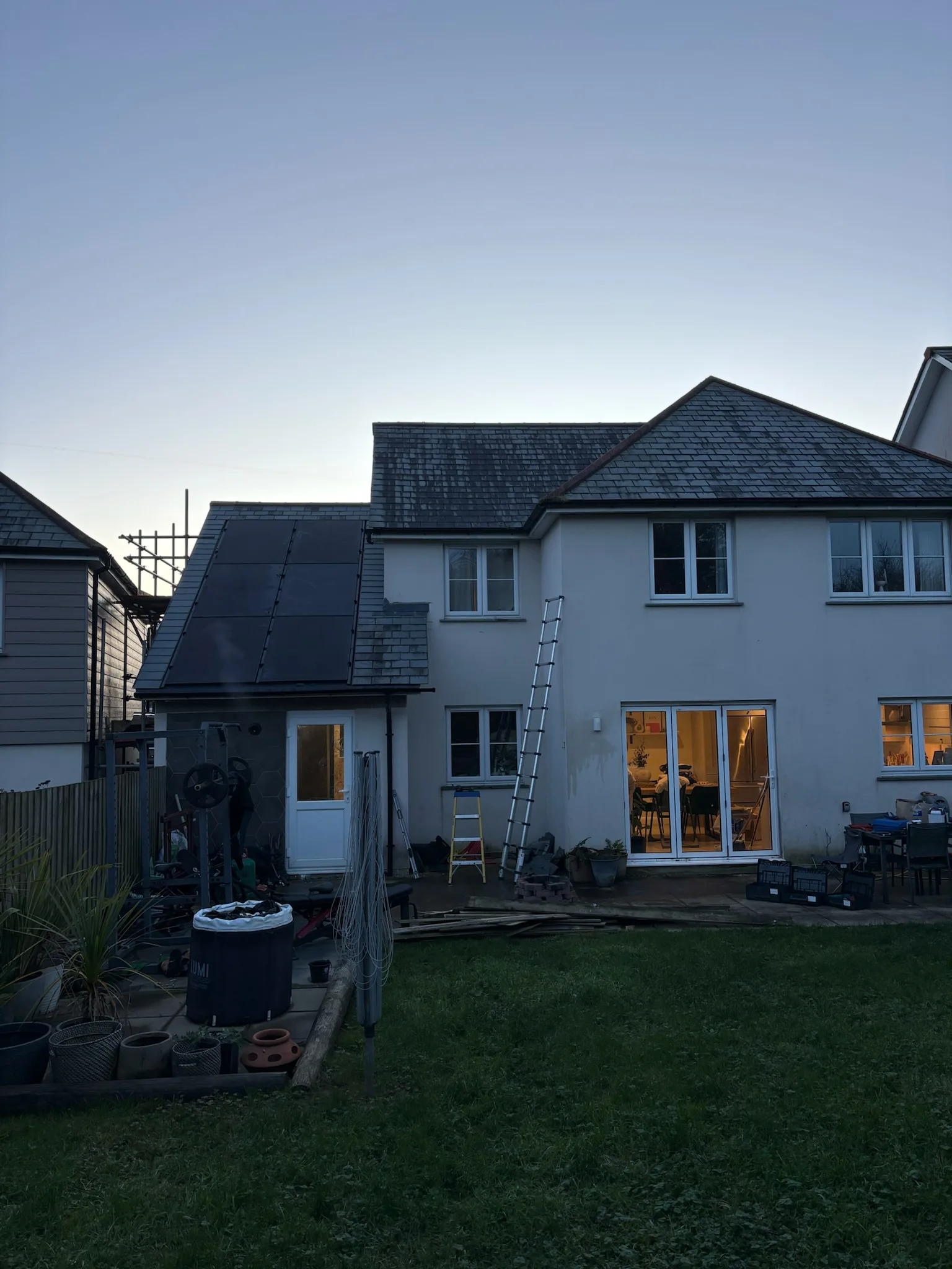 Rear view of a two-storey house and garden with dark panels on a single-storey pitched roof, two ladders against the wall and scaffolding at the side.