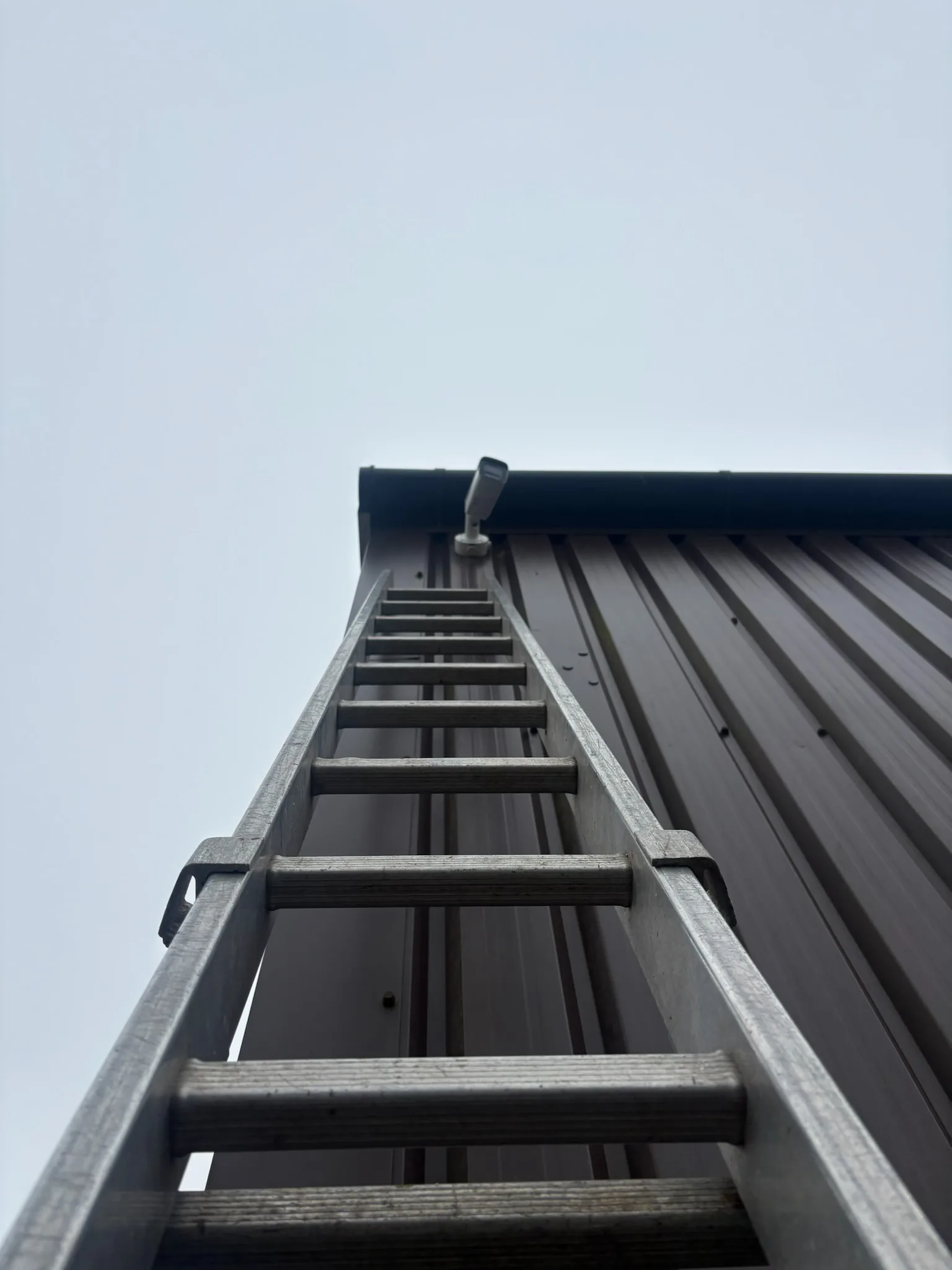 View looking upwards along a metal access ladder against dark corrugated wall cladding beneath an overcast sky.