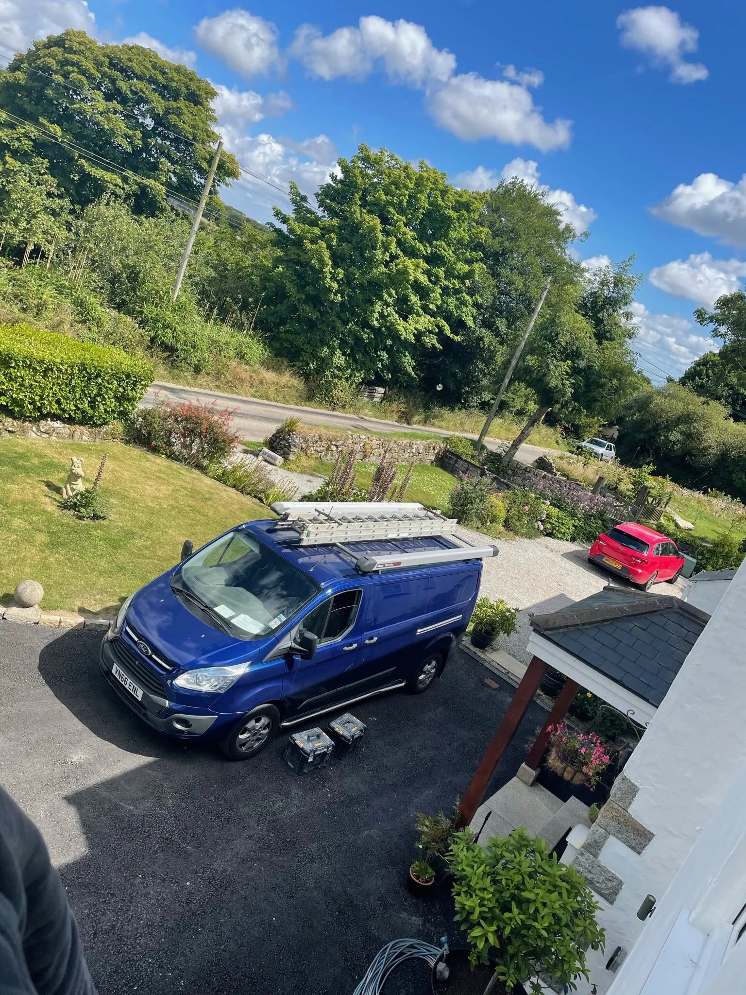 Elevated view of a blue work van with a ladder secured to its roof, parked on a driveway beside tools and a house.