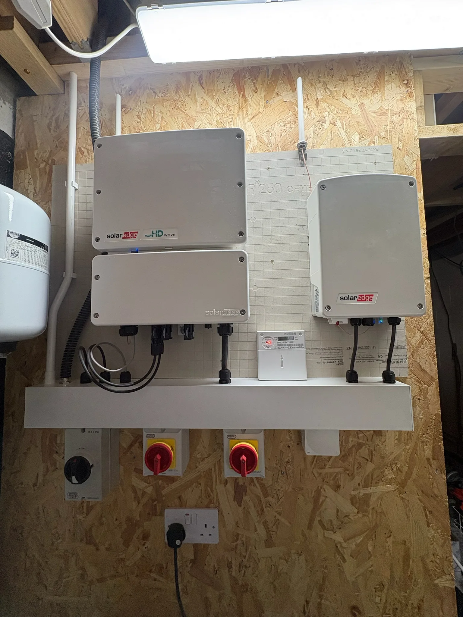 Three grey SolarEdge units mounted on a white panel above isolator switches and a socket in a timber-walled room.