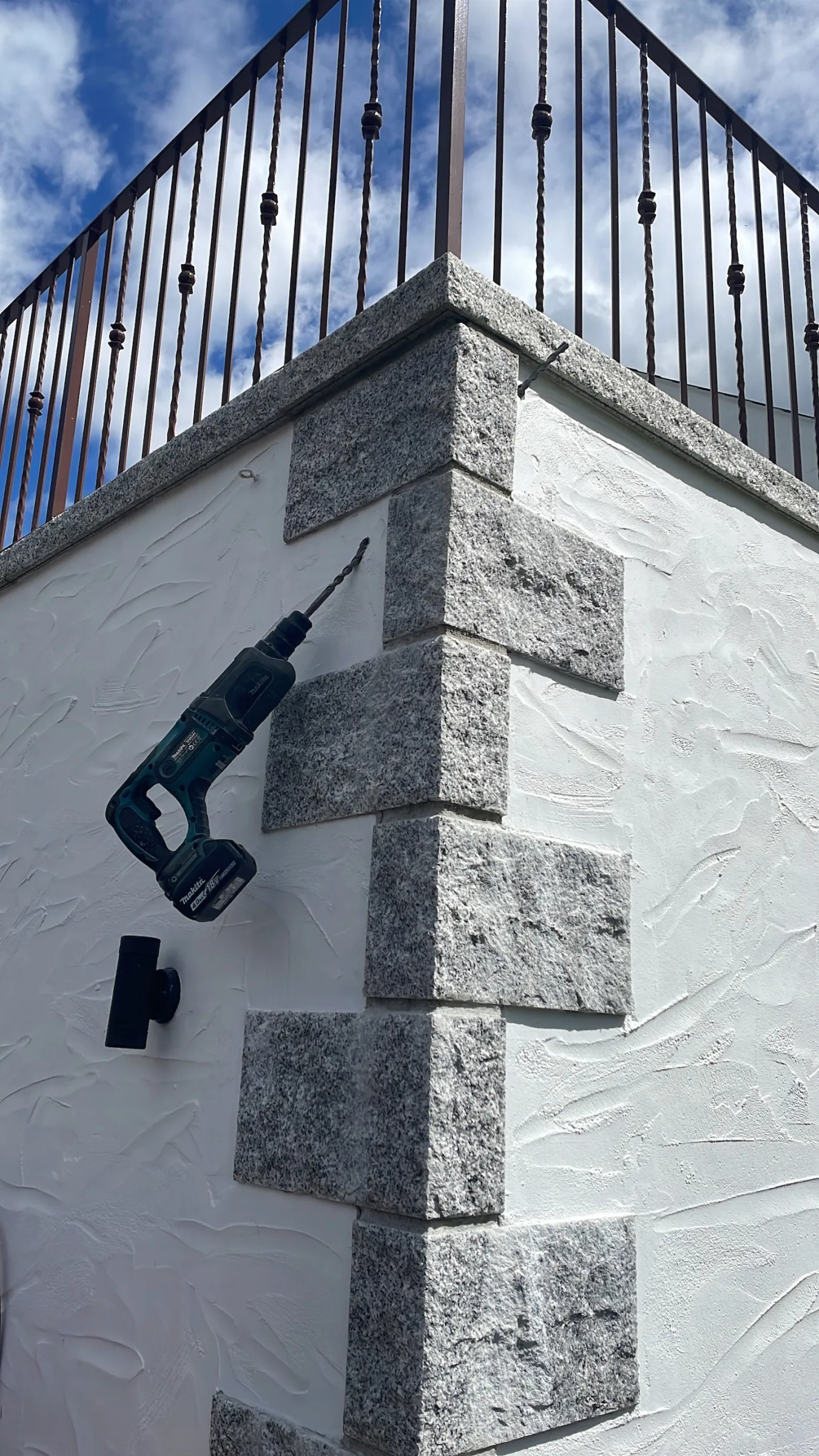 Cordless drill with a long bit positioned against the grey corner blocks of a white rendered wall beneath black metal railings.