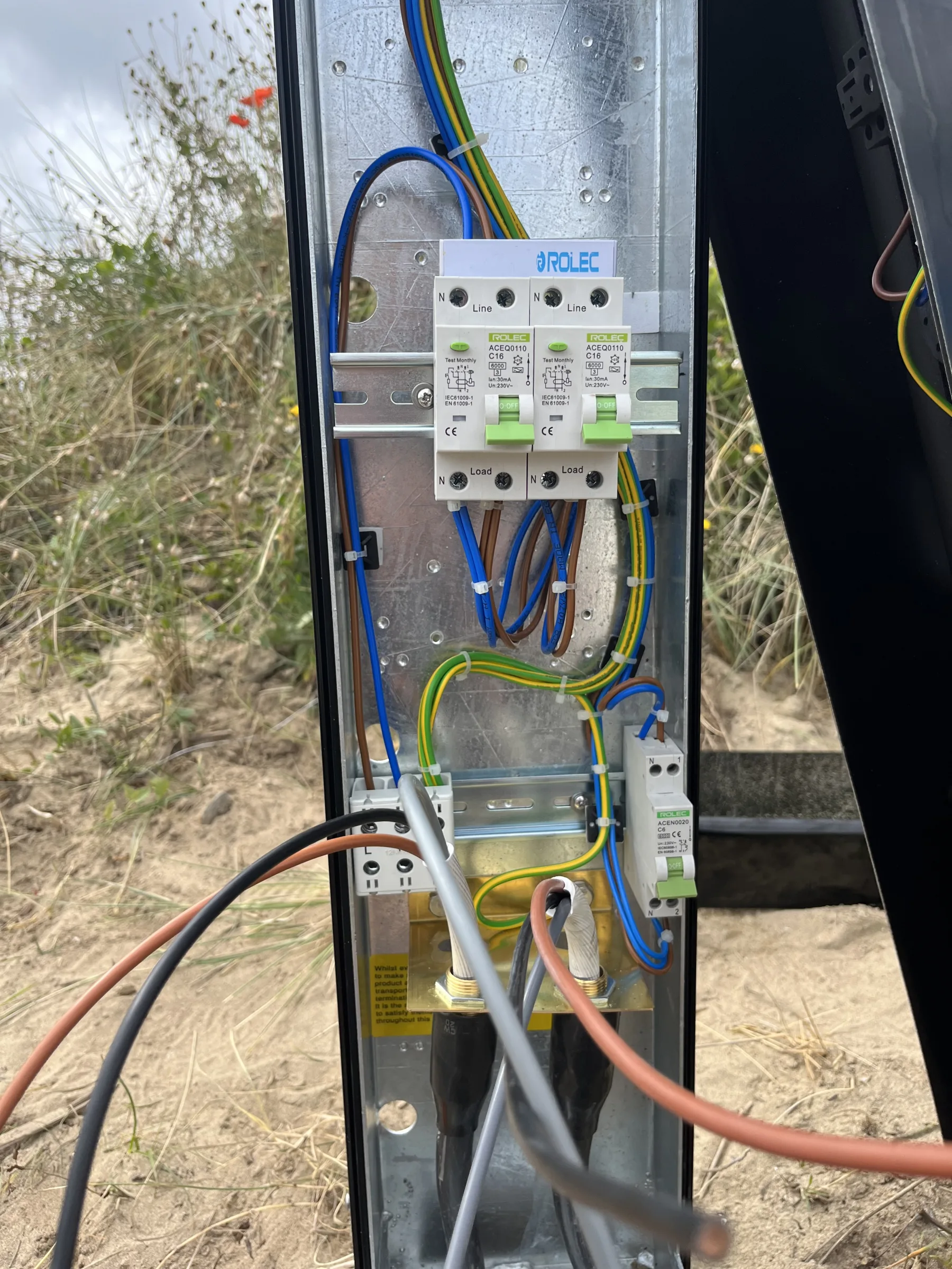 Open electrical post showing a metal backplate, rail-mounted electrical devices, blue and brown wiring, green-and-yellow earth wires and incoming cables.