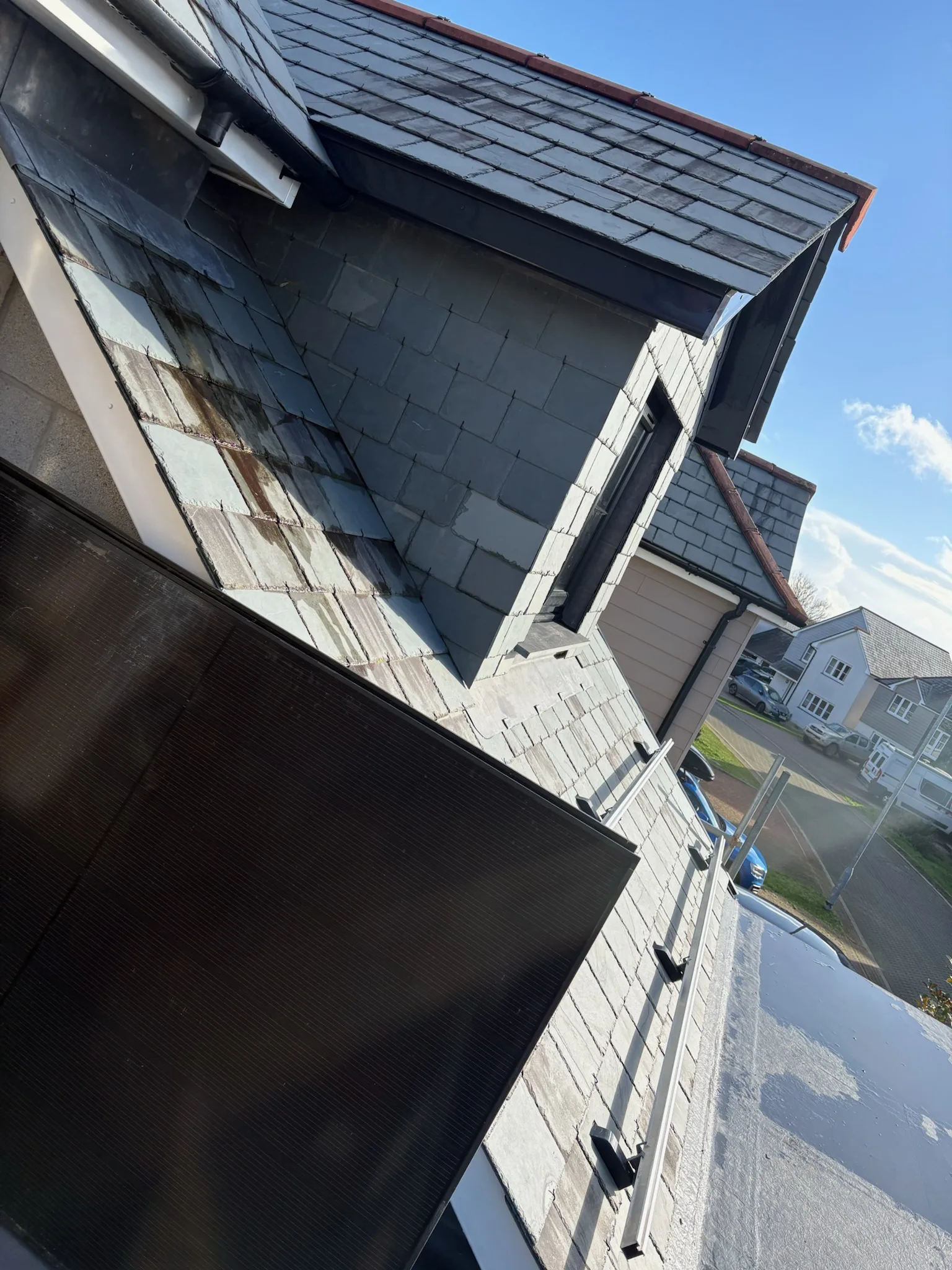 Dark solar panel fitted on a sloping roof beside a slate-clad dormer under a blue sky.
