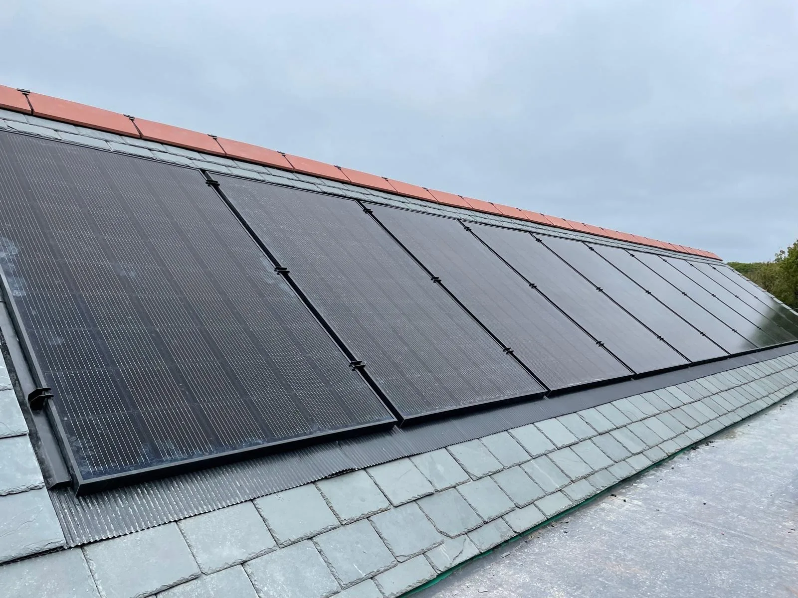 Black solar panels fitted in a continuous row across a pitched grey slate roof with red ridge tiles.